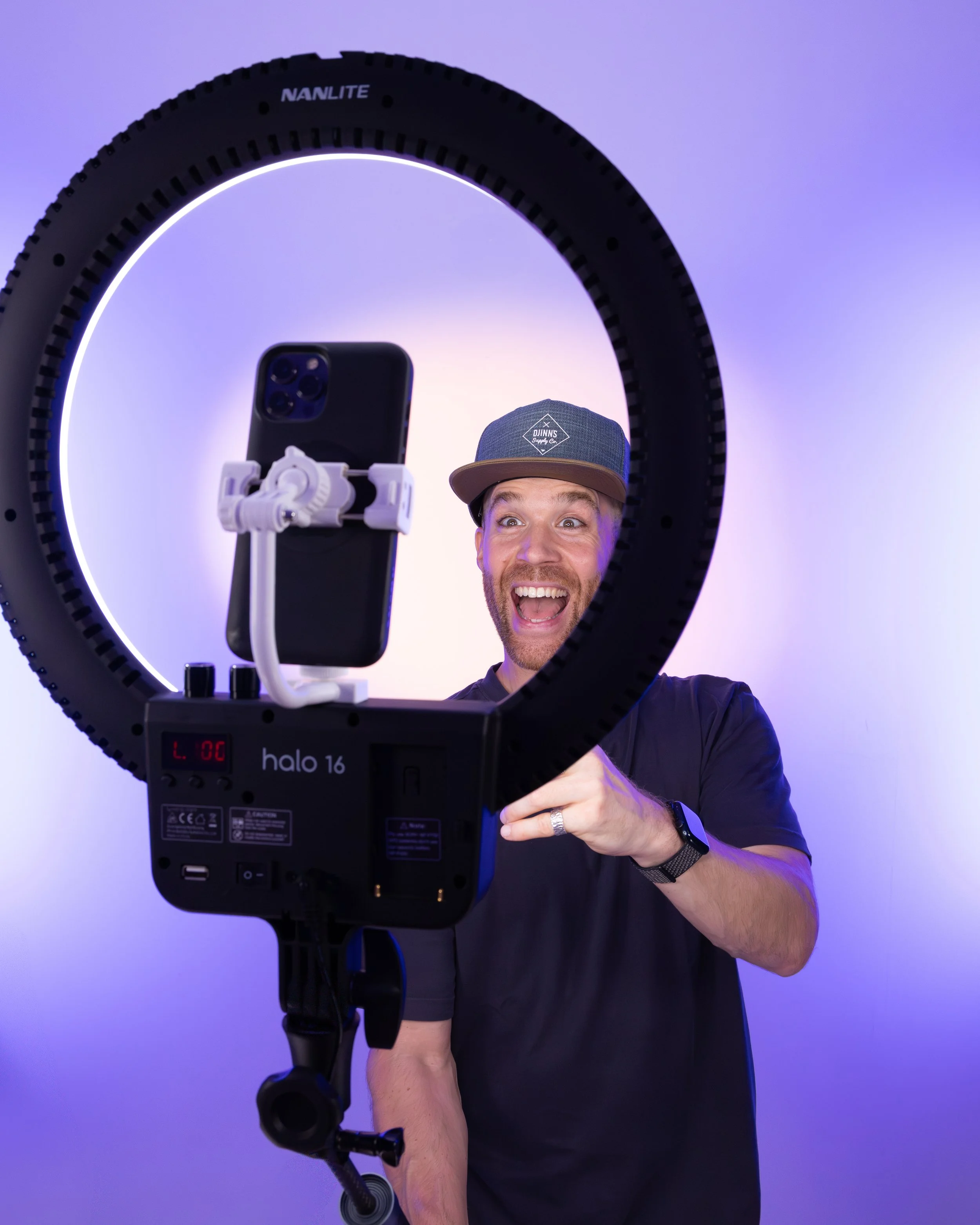 Erik van der Ven in a black shirt wearing a baseball cap taking a selfie with a smartphone mounted on a tripod, seen through a ring light against a purple background.