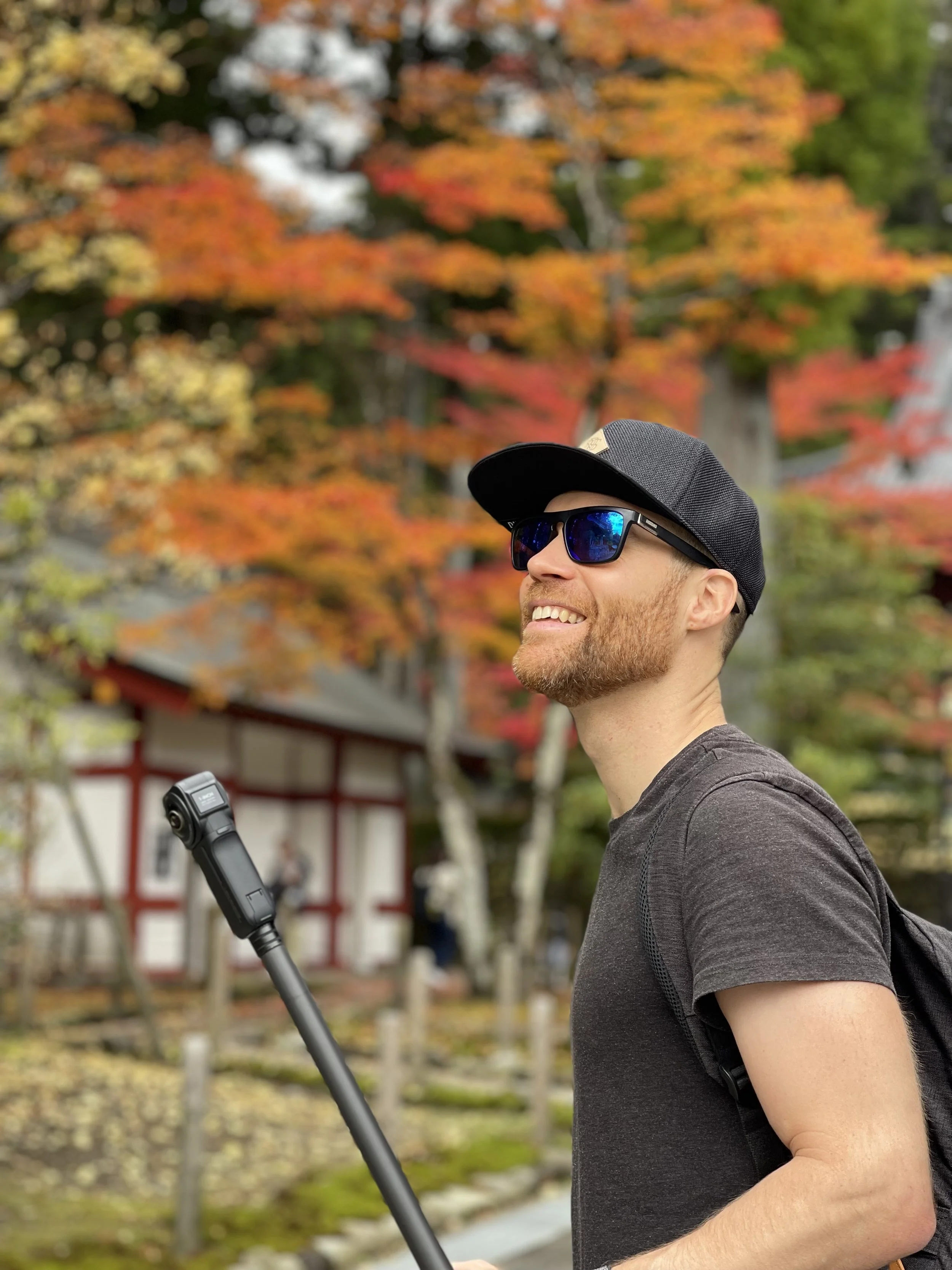 Erik van der Ven with a beard, wearing sunglasses, a black cap, and a dark t-shirt, smiling outdoors during fall in Japan. He's holding a selfie stick, with a background of colorful autumn trees and traditional Japanese architecture.