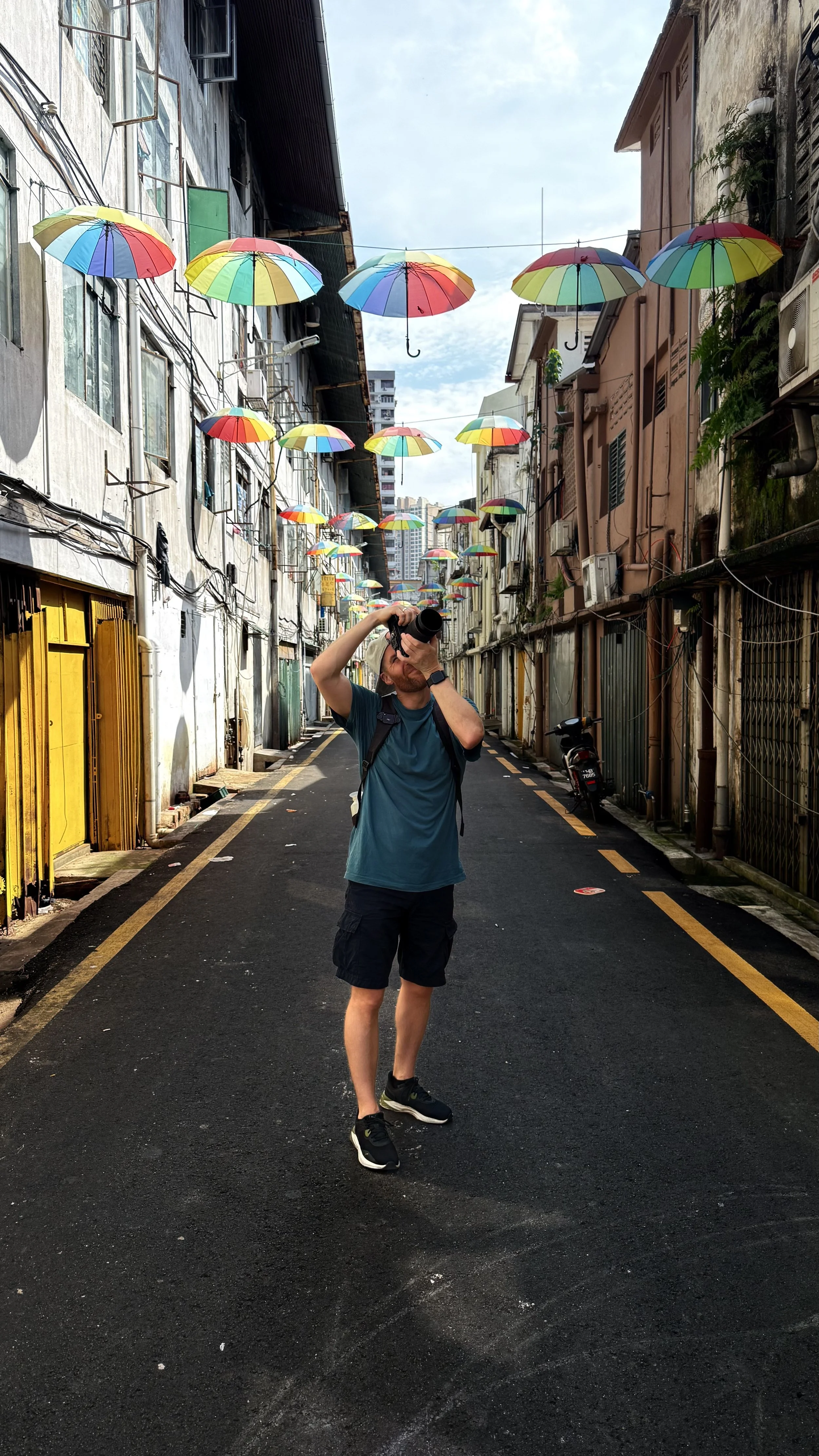 Erik van der Ven standing in an alleyway decorated with colorful umbrellas hanging overhead, with buildings on both sides and a city skyline in the background.