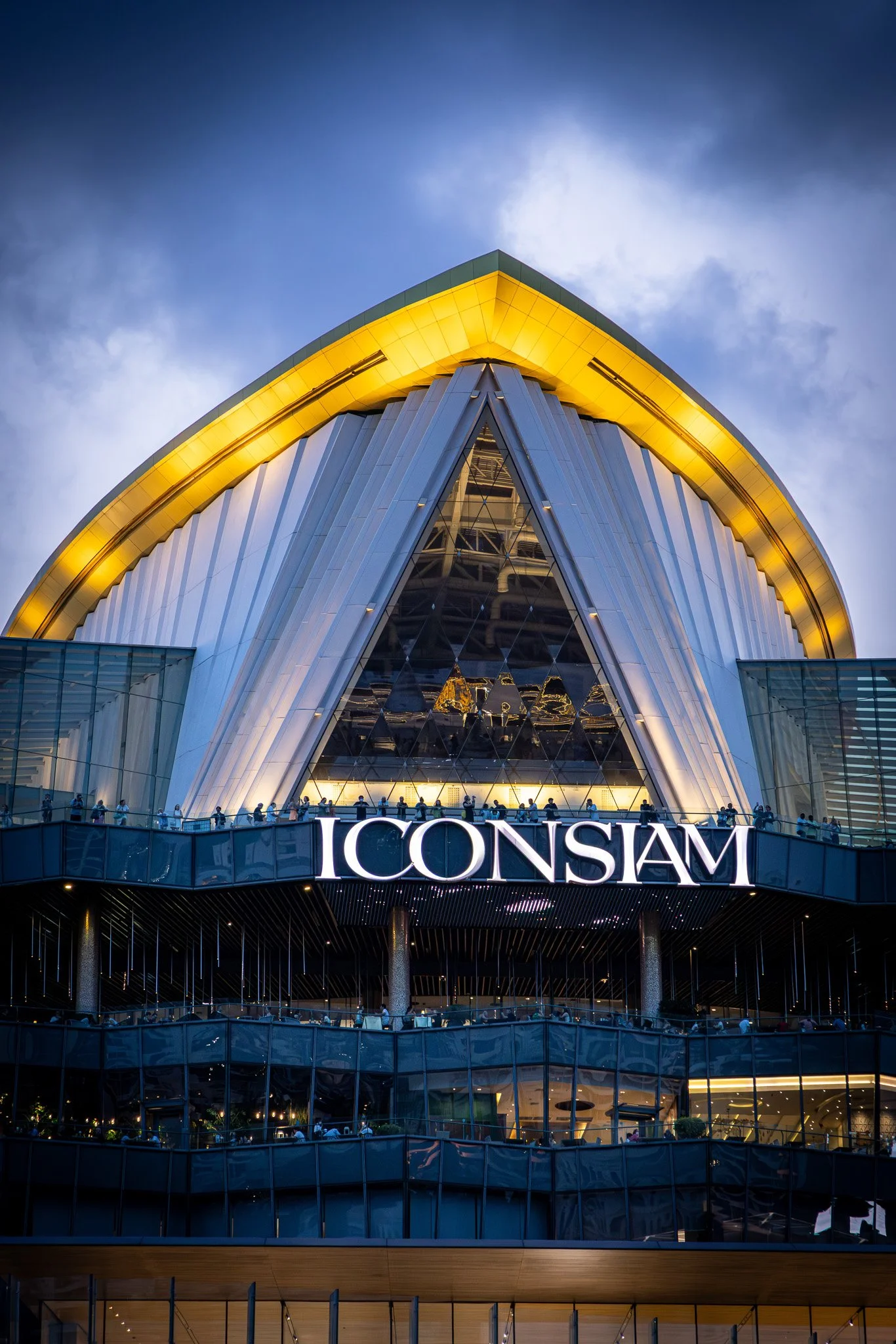 A modern building with a curved roof illuminated in yellow lights, featuring reflective glass panels and multiple floors with people visible inside, and a sign reading 'ICONSIAM' at the front.