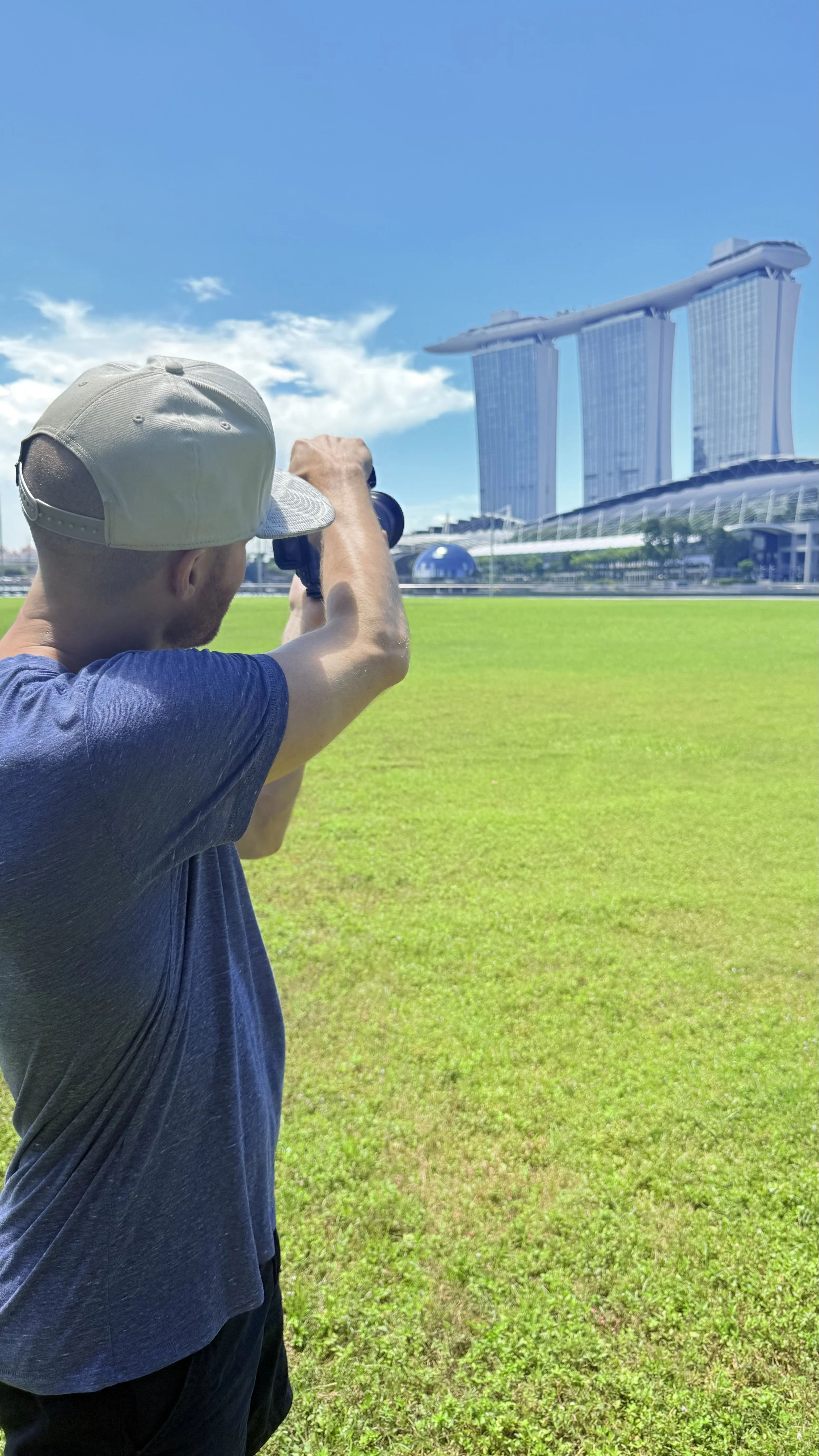 Erik van der Ven in a white cap and dark T-shirt is taking a photo with his camera on a lush green field. In the background, there is the Marina Bay Sands hotel with its three towers and rooftop structure, under a bright blue sky with some clouds.