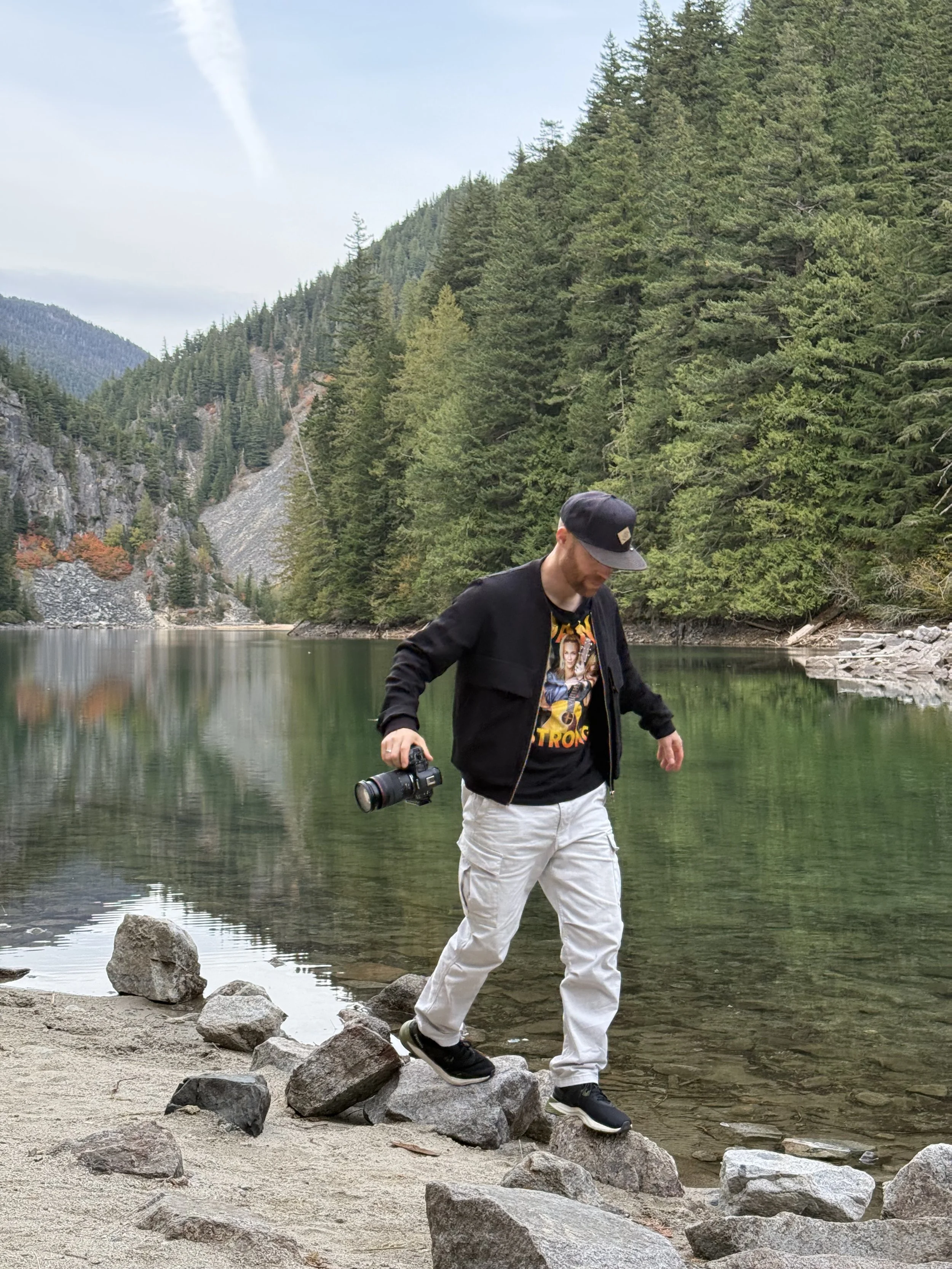 Erik van der Ven wearing a black cap, black jacket, and white pants walking on rocks by a lake with green trees and mountains in the background.