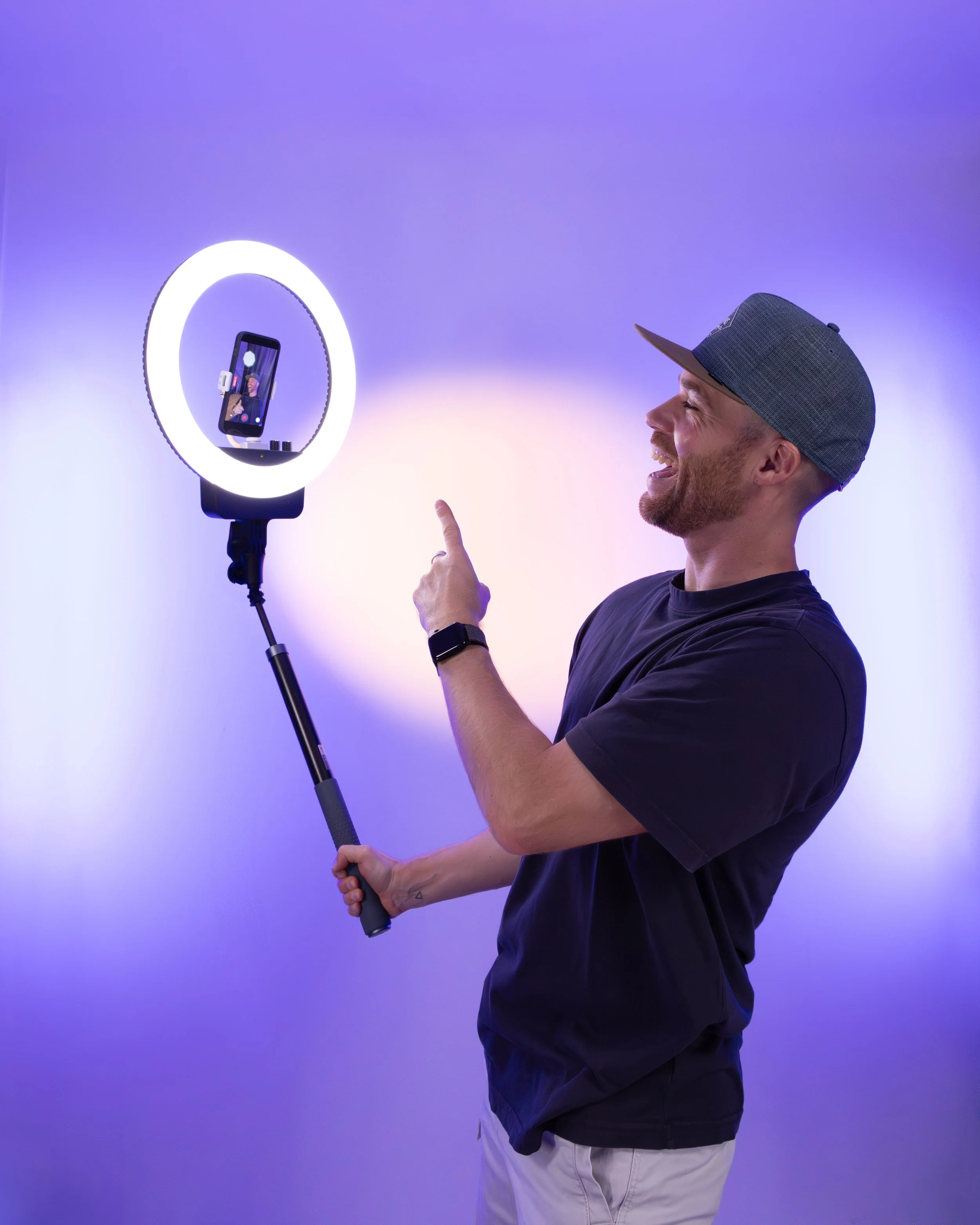 Erik van der Ven with a beard, wearing a gray baseball cap, a dark T-shirt, and light-colored pants, is smiling and pointing at a smartphone mounted on a selfie ring light stand.