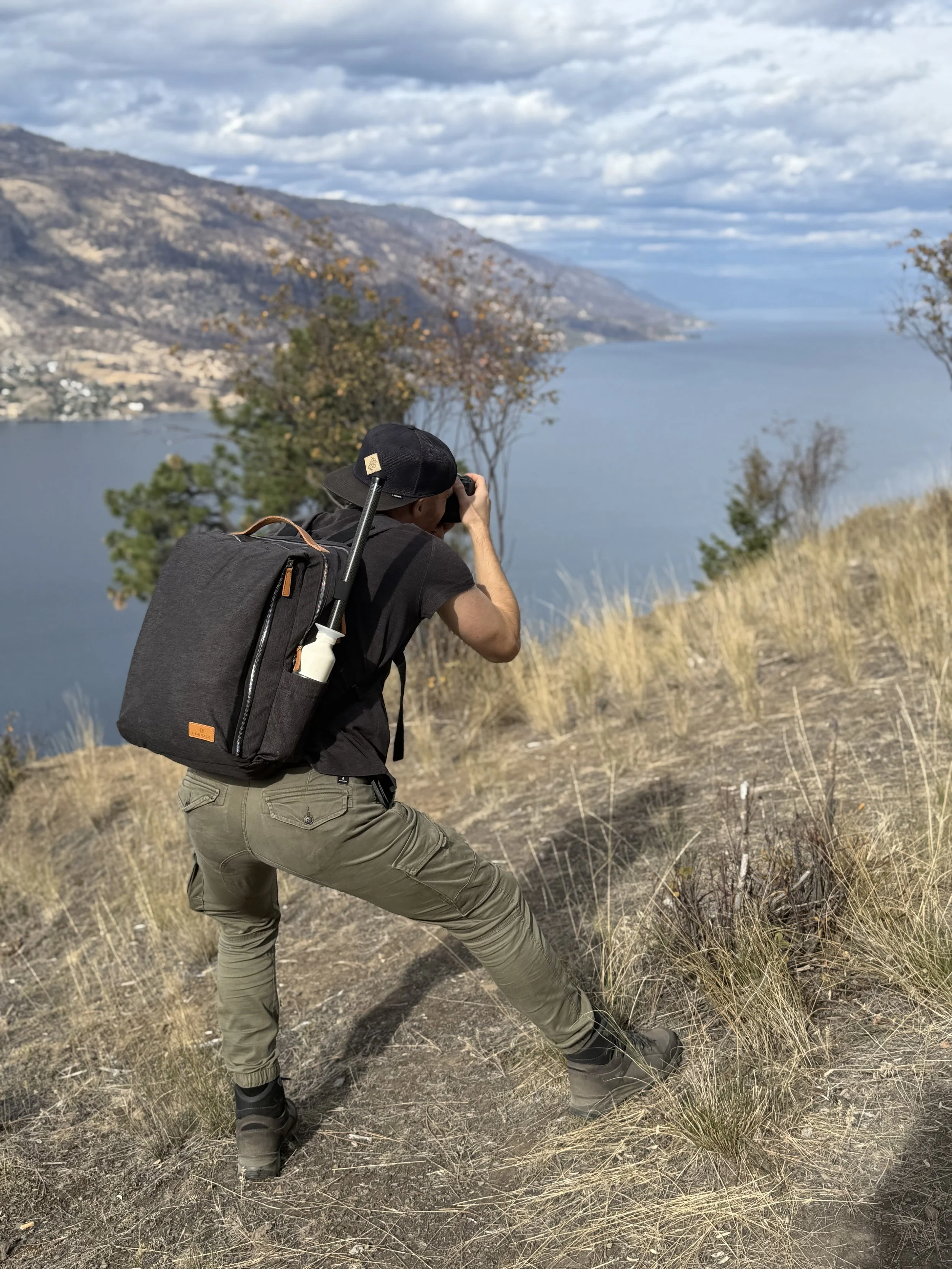 Erik van der Ven with a black hat, backpack, and hiking boots taking a photograph by a lake with mountains in the background, and dry grass in the foreground.