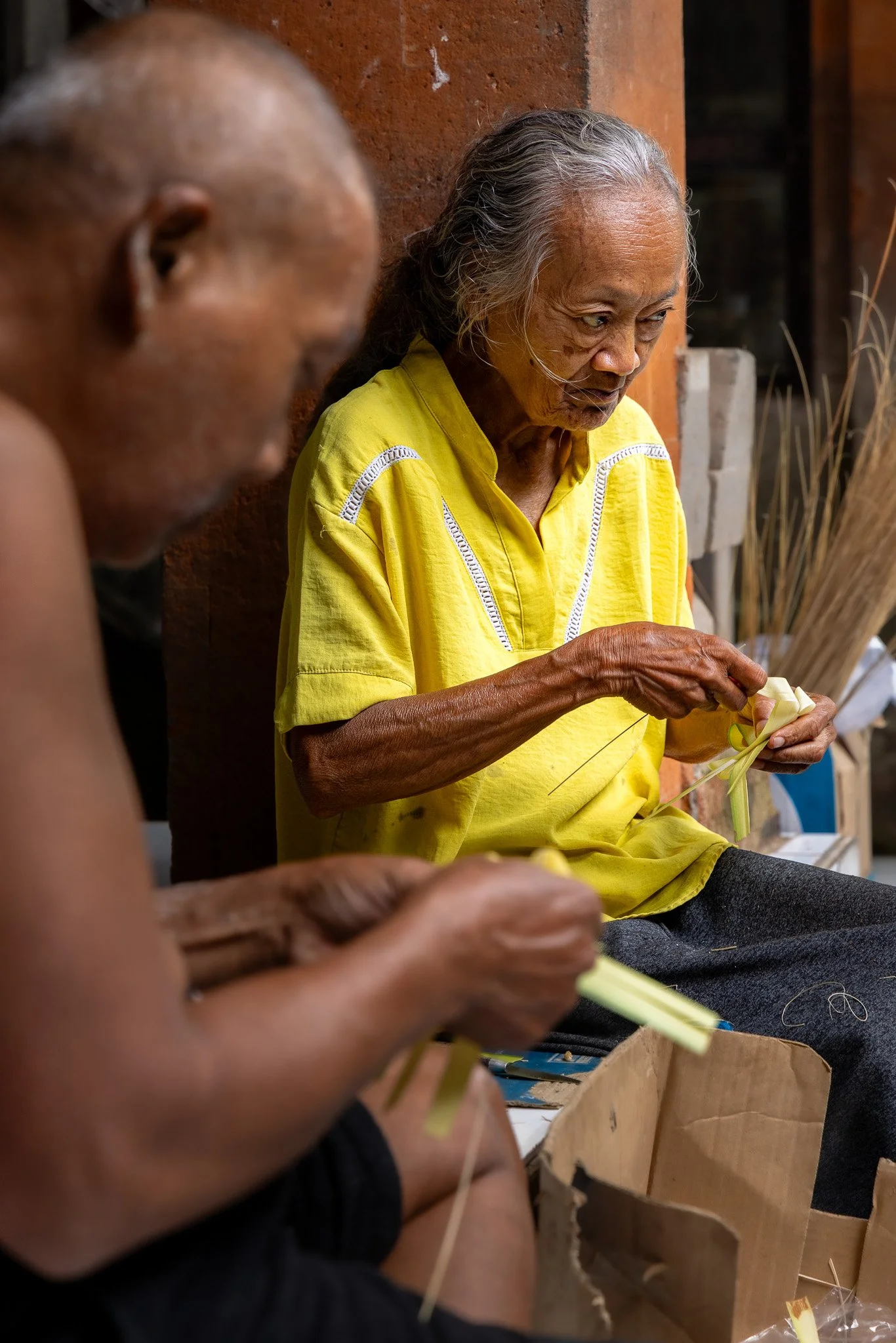 Two elderly people with gray hair engaging in traditional craft work, sitting against a brick wall, with one in a yellow shirt focusing on the craft, and the other partially visible in the foreground.
