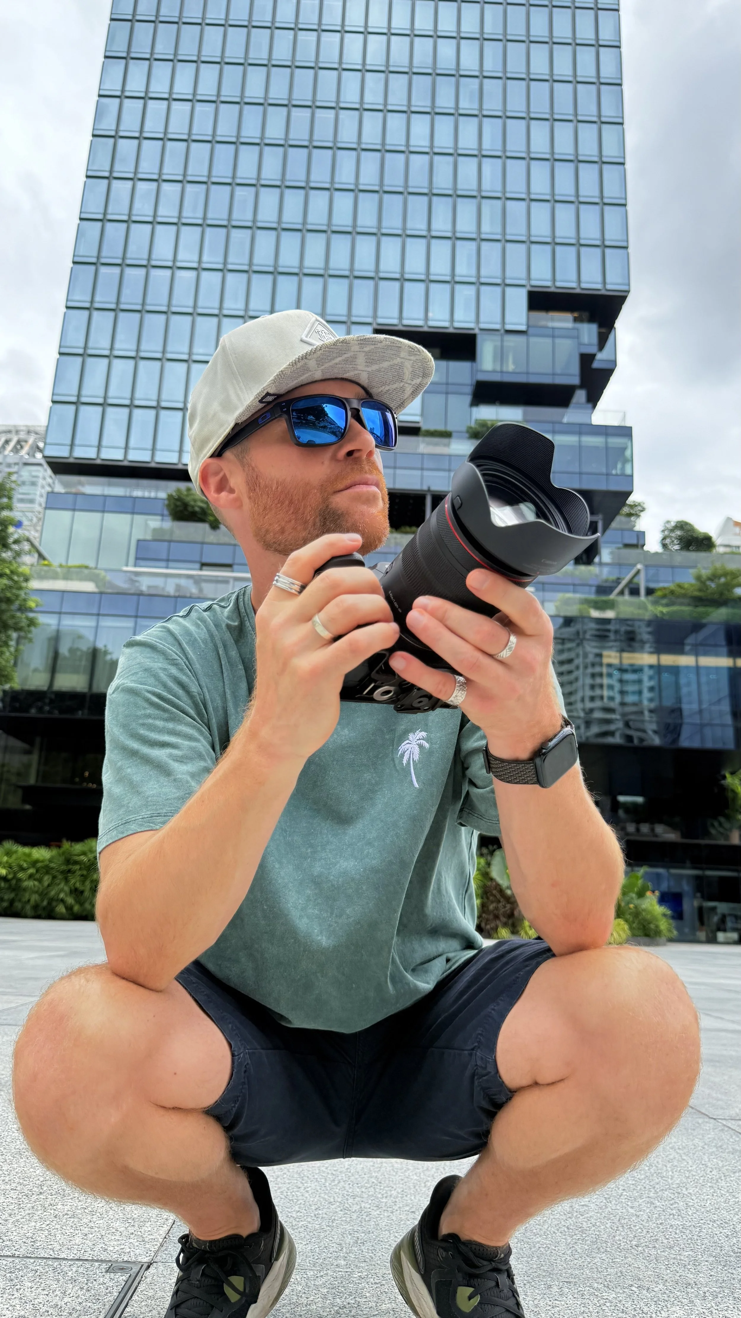 Erik van der Ven squatting outdoors in front of a tall modern glass building, holding a professional camera with a large lens, wearing sunglasses, a cap, a green t-shirt, black shorts, a watch, and multiple rings.