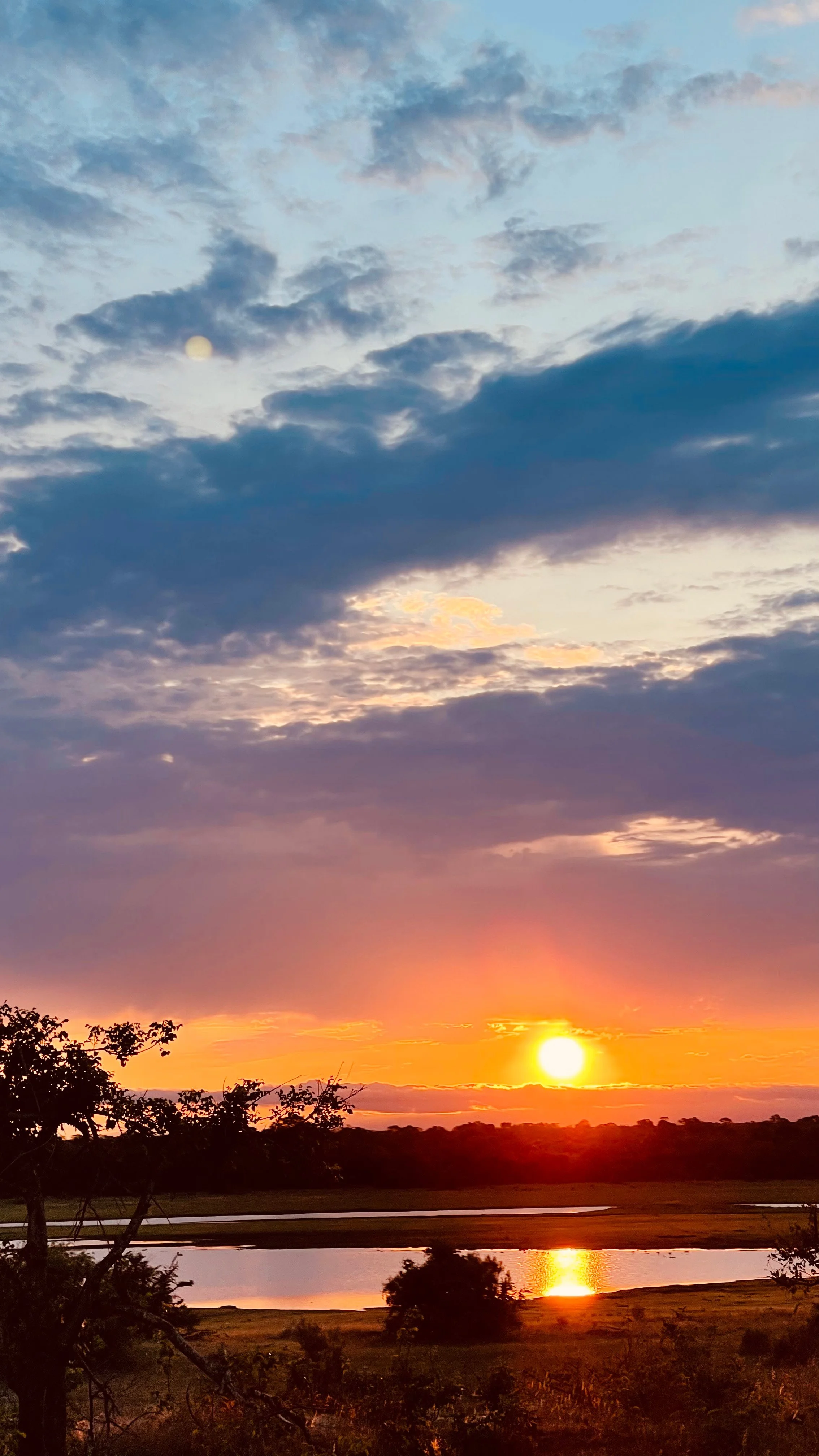 A sunset over a lake with trees in the foreground and clouds in the sky.