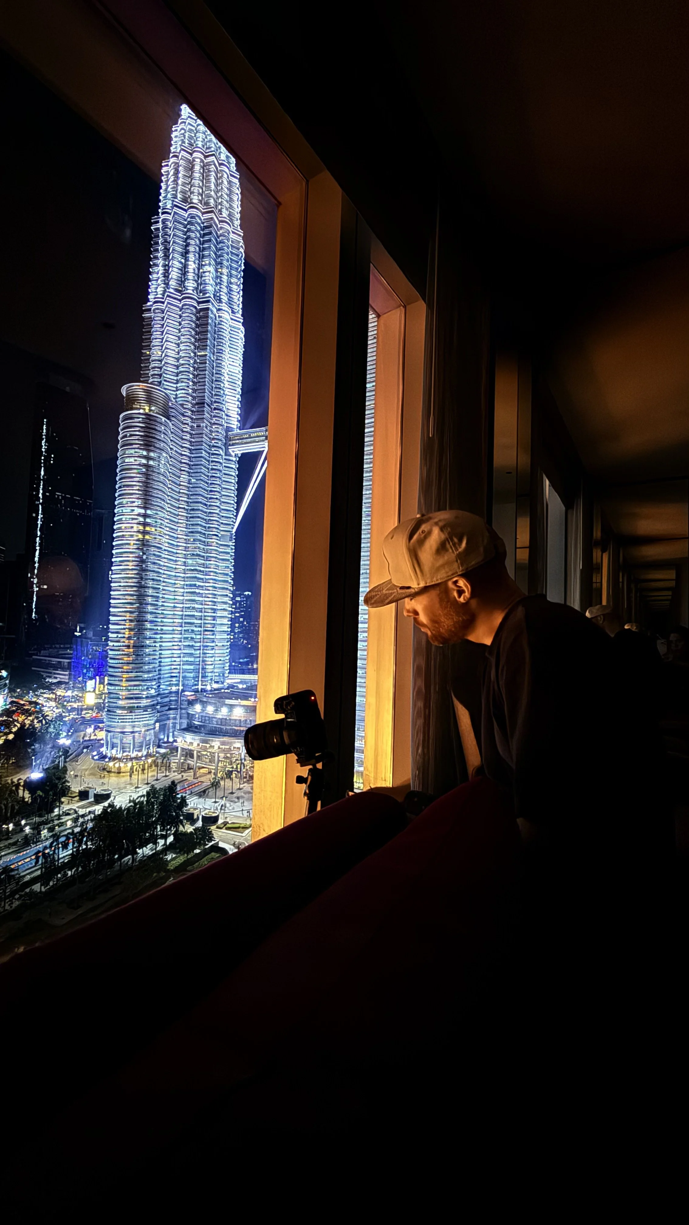 Erik van der Ven wearing a beige baseball cap and black shirt sitting by a window observing the illuminated Petronas Towers at night in Kuala Lumpur, Malaysia.