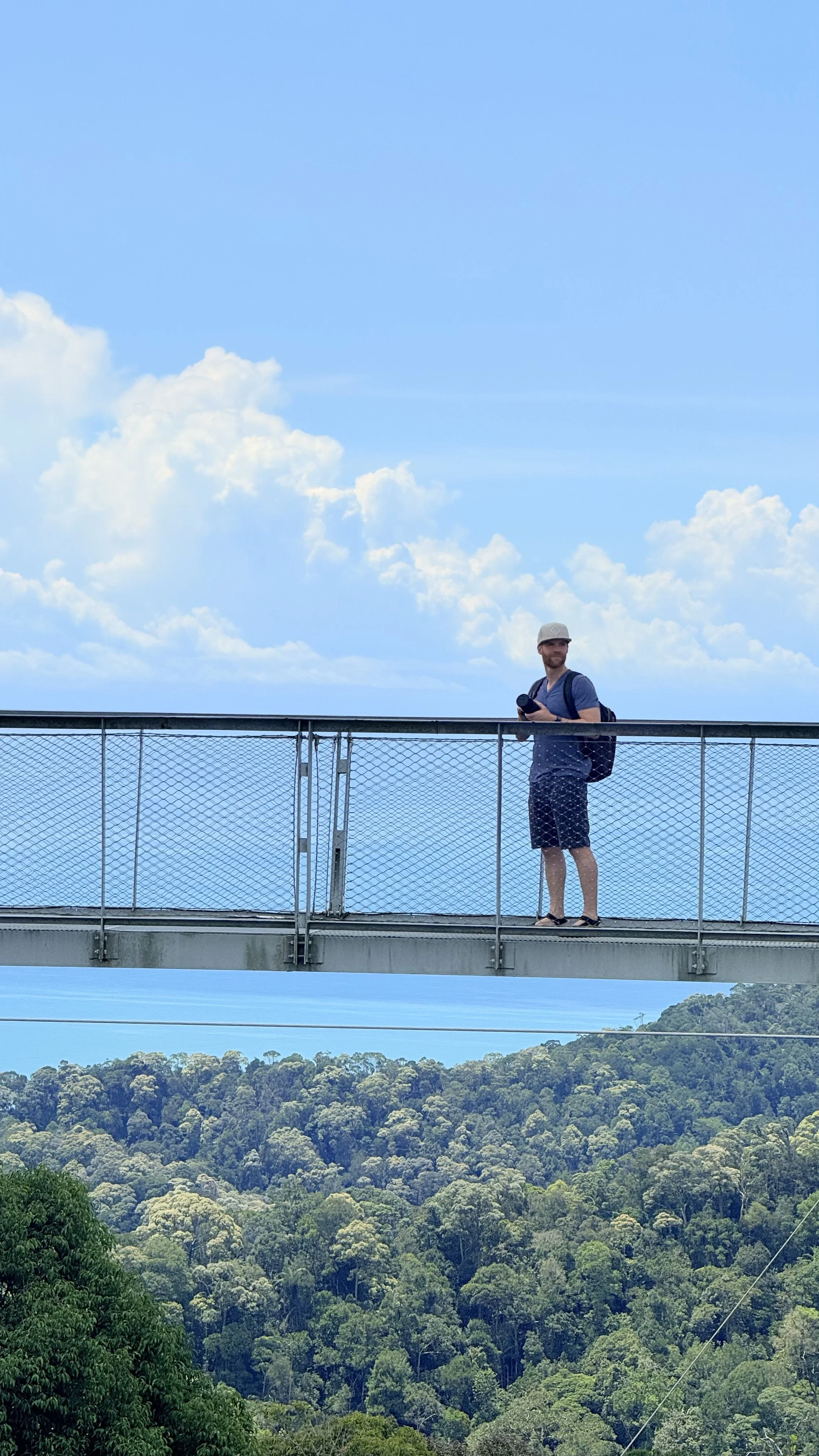 Erik van der Ven standing on a bridge, wearing a gray cap, blue shirt, dark shorts, and a backpack, with a scenic landscape of a forested area and a partly cloudy sky in the background.