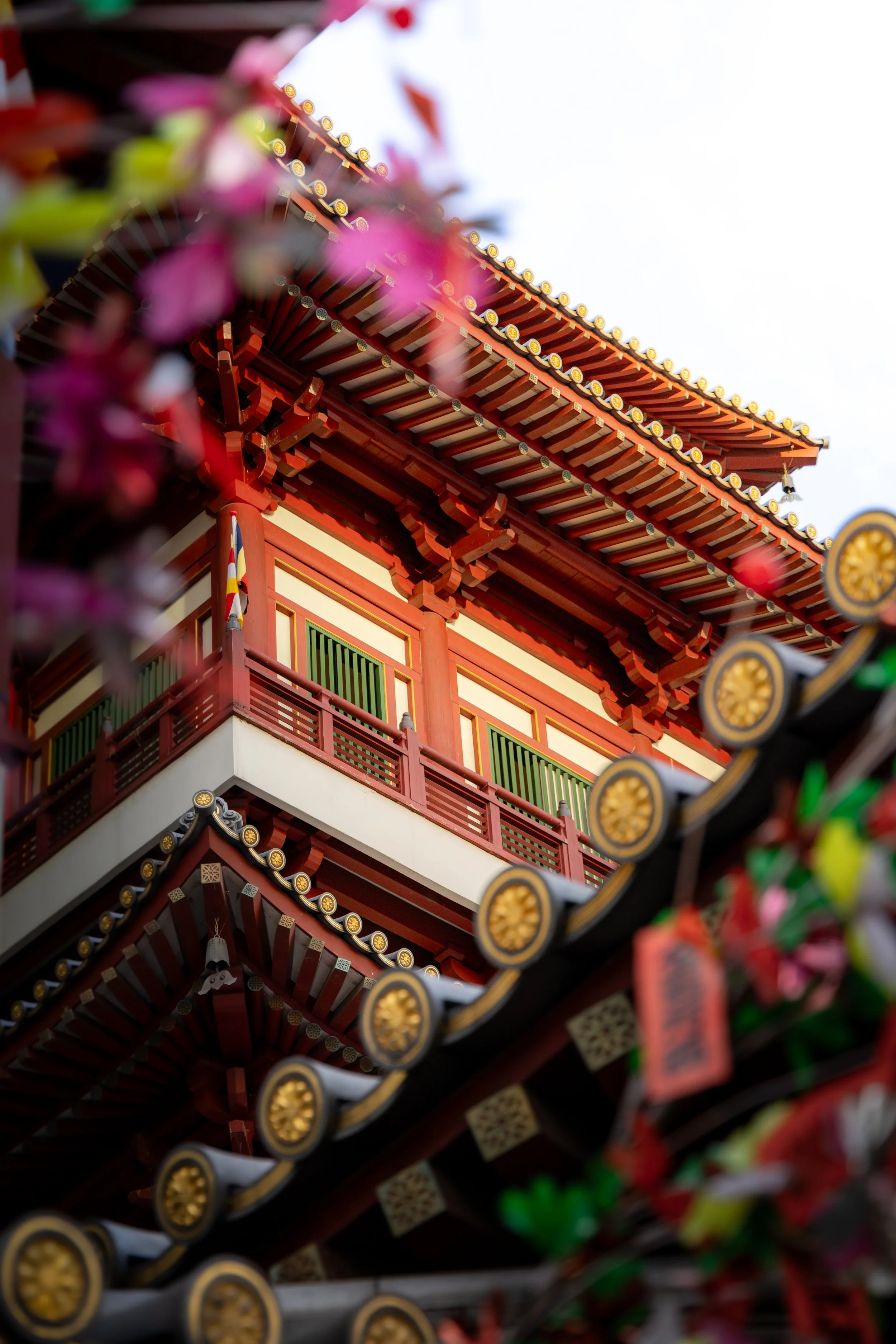 A traditional Asian building with ornate red and gold architectural details, multi-tiered roof with decorative ceramic tiles, and colorful hanging lanterns, partially obscured by pink flowers in the foreground.
