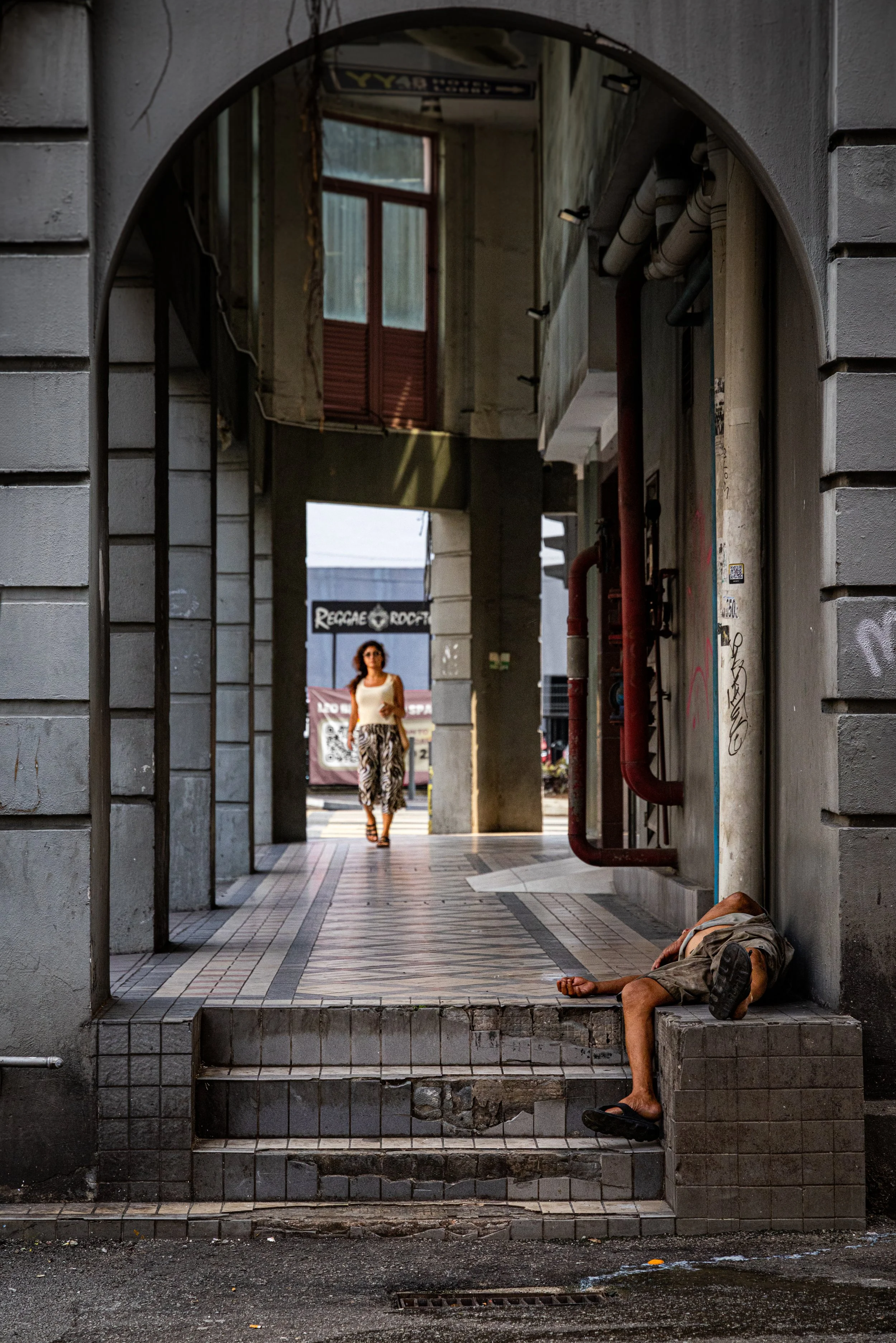 A street scene with a person lying on the ground near the corner of a building and a woman walking in the background. The person on the ground appears to be a homeless individual, and the scene is framed by an archway.