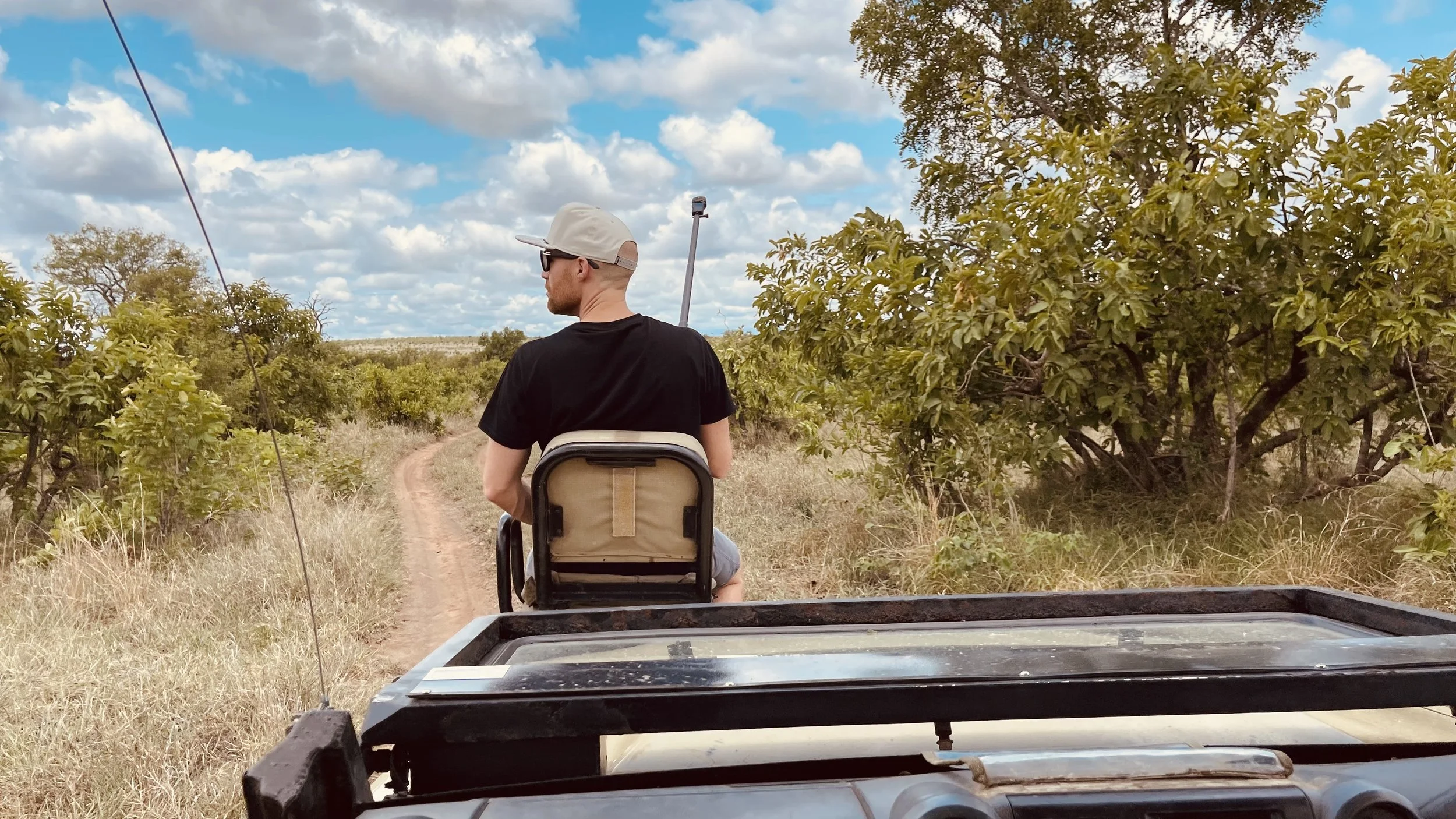 Erik van der Ven wearing a white cap and sunglasses sitting in front of a safari truck on a dirt path through a rural area with trees and grass under a partly cloudy sky.