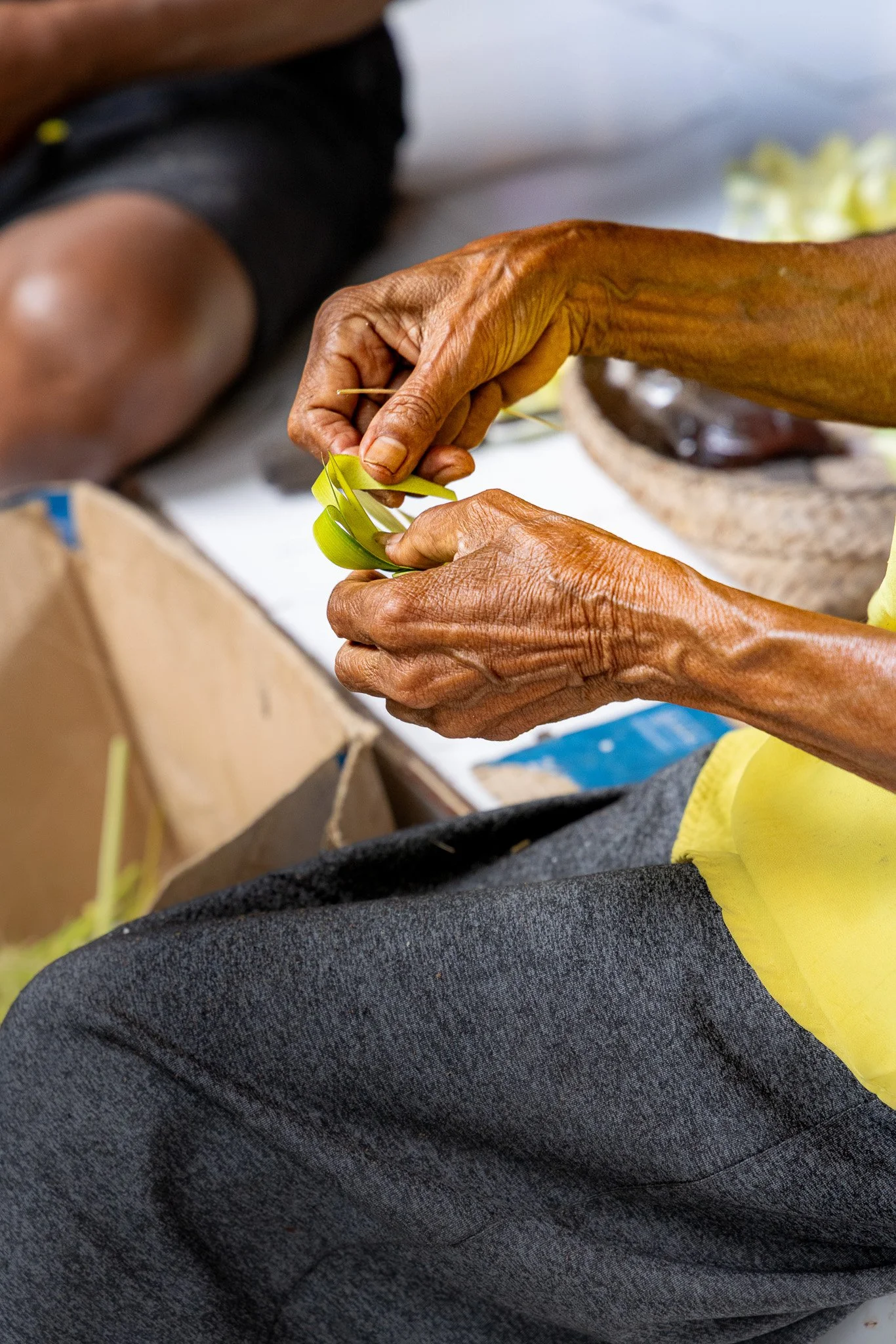 Close-up of elderly person's hands weaving a bright yellow-green leaf or plant material, with a blurred background showing part of their arm, clothing, and a box.