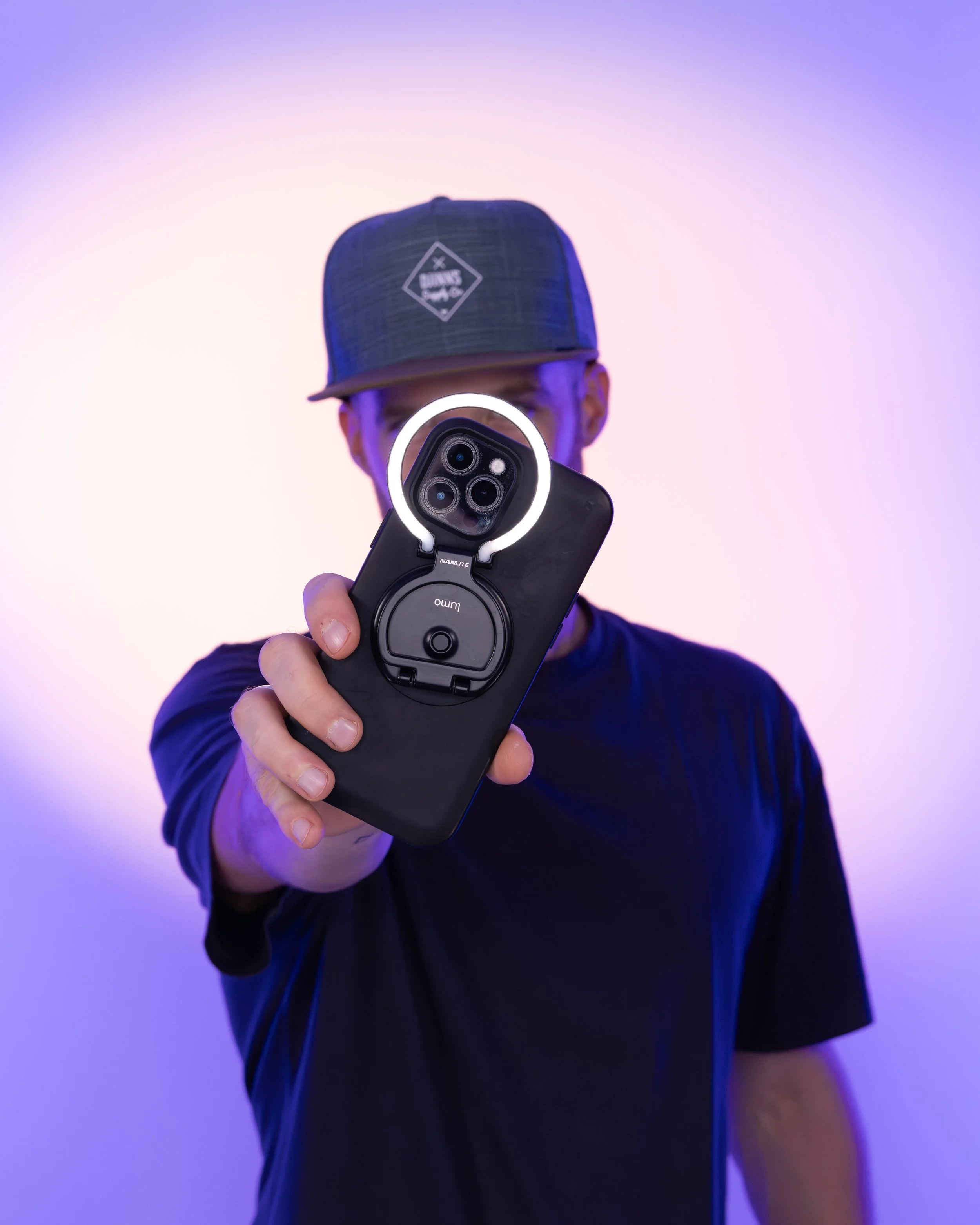 Erik van der Ven holding a smartphone with a ring light attached, wearing a blue cap and shirt, against a purple and pink gradient background.