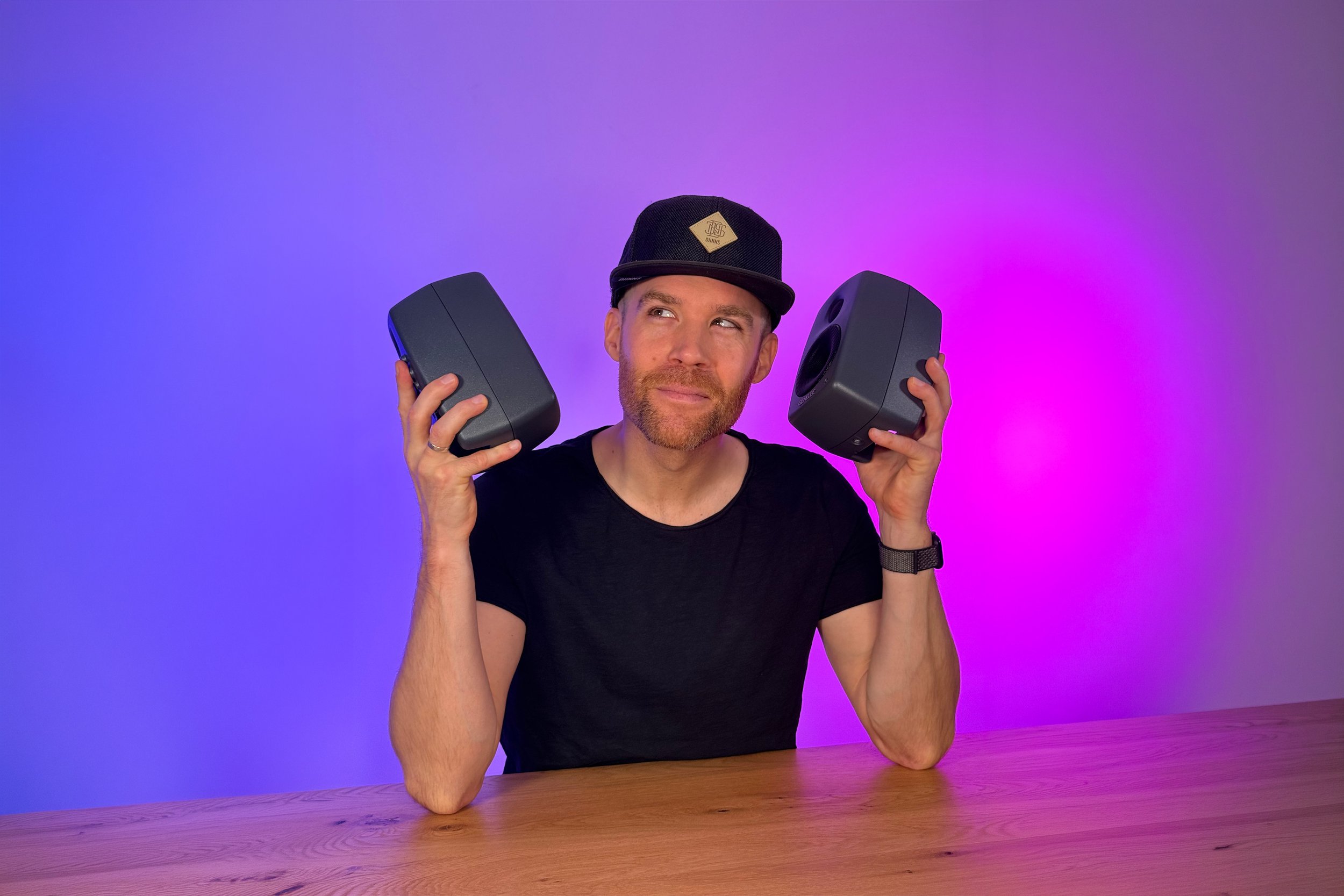 Erik van der Ven with a beard and wearing a black T-shirt and a black cap with a logo, holding two speakers near his ears, sitting at a wooden table with a purple and pink gradient background.