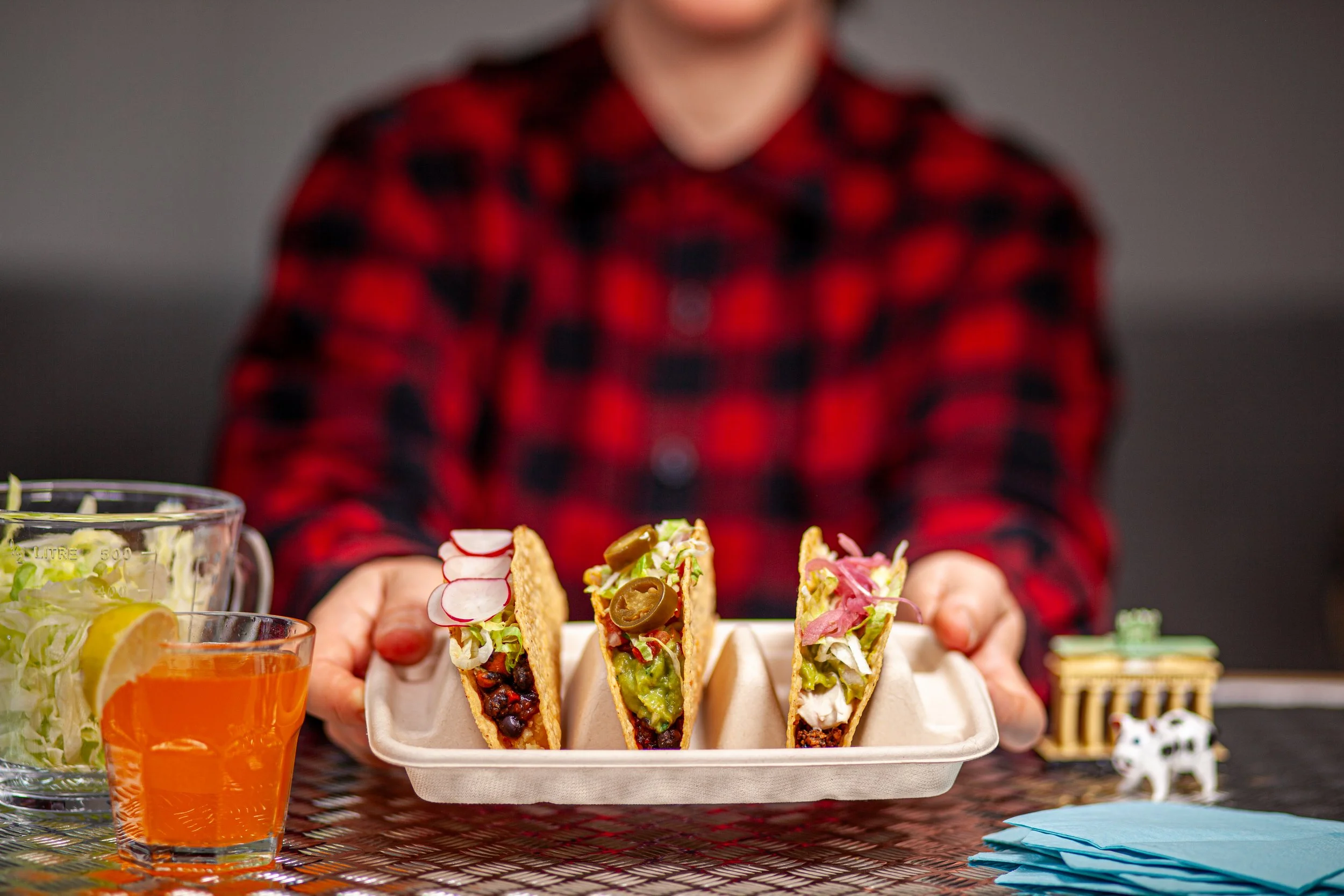 A person in a red flannel shirt holds up a plate with three vegan tacos in crispy shells with various toppings..