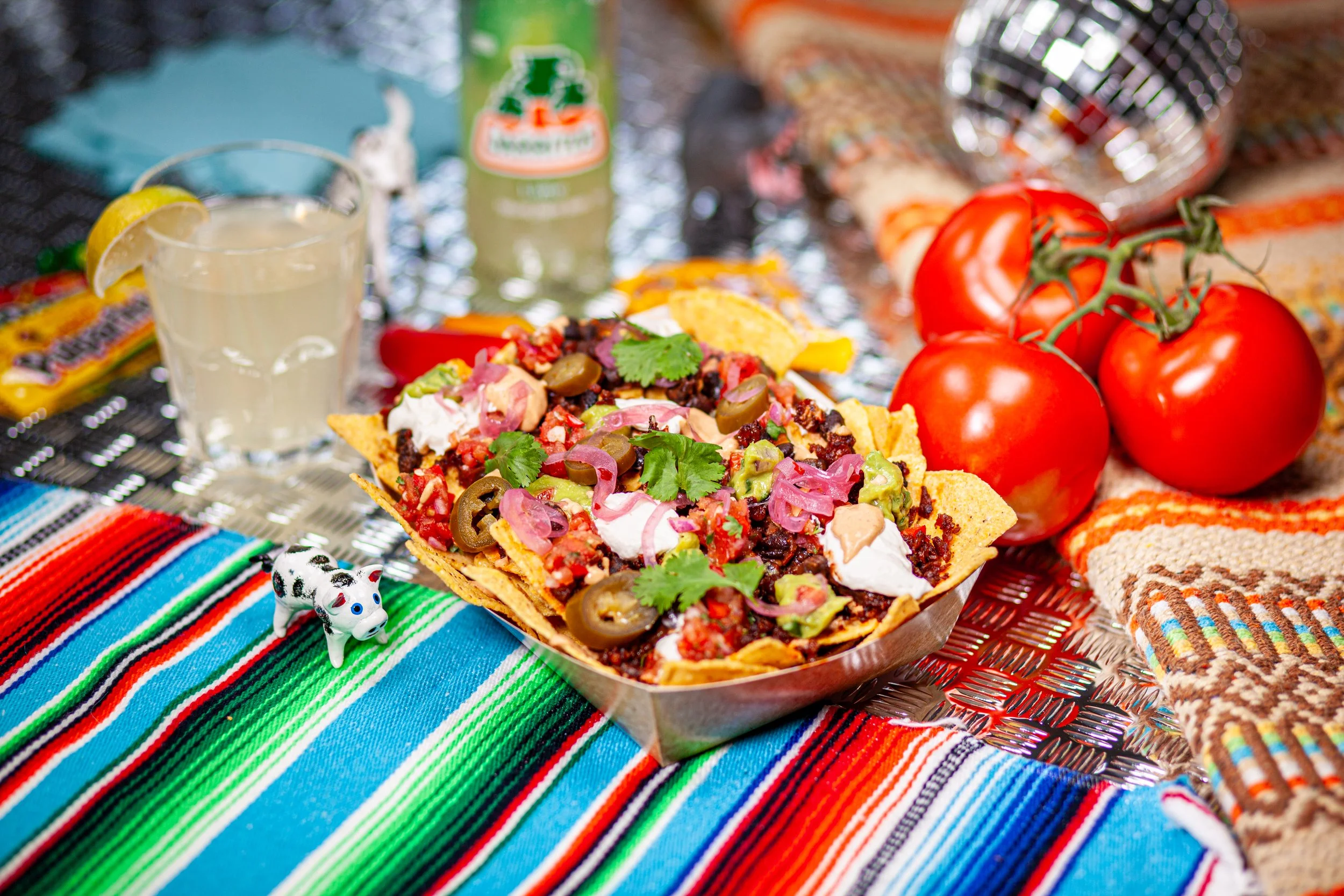 A plate full of nachos piled with vegan toppings on a striped cloth with a small black and whit pig figuring on the left and a bottle of soda in the background.