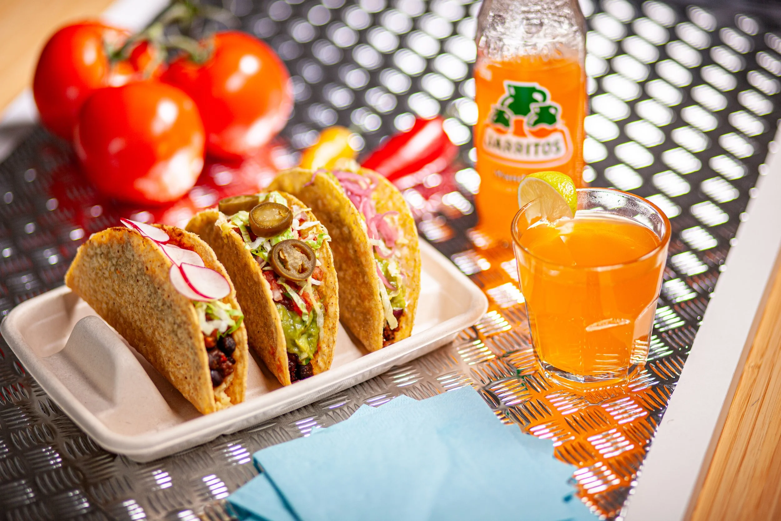 Three vegan tacos with various fillings and toppings on a metal surface with tomatoes and chilis in the background/