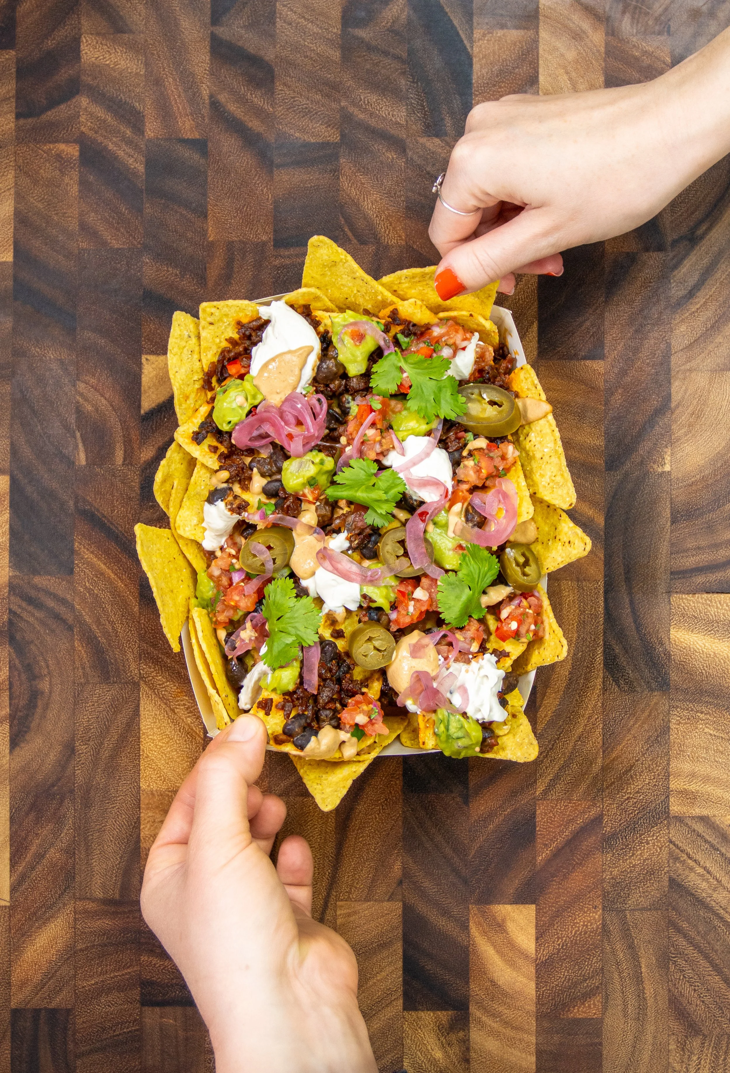 Two people sharing a large plate of loaded nachos topped with seasoned tofu, black beans, tomato salsa, sliced jalapeños, pickled red onions, guacamole, vegan sour cream, vegan cheese sauce, and cilantro, placed on a dark wooden table.