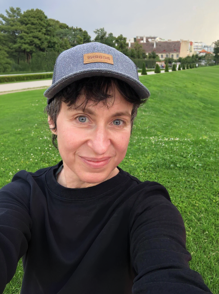 A person wearing a gray cap and a black long-sleeve shirt, smiling while taking a selfie outdoors in a grassy park with trees and buildings in the background.