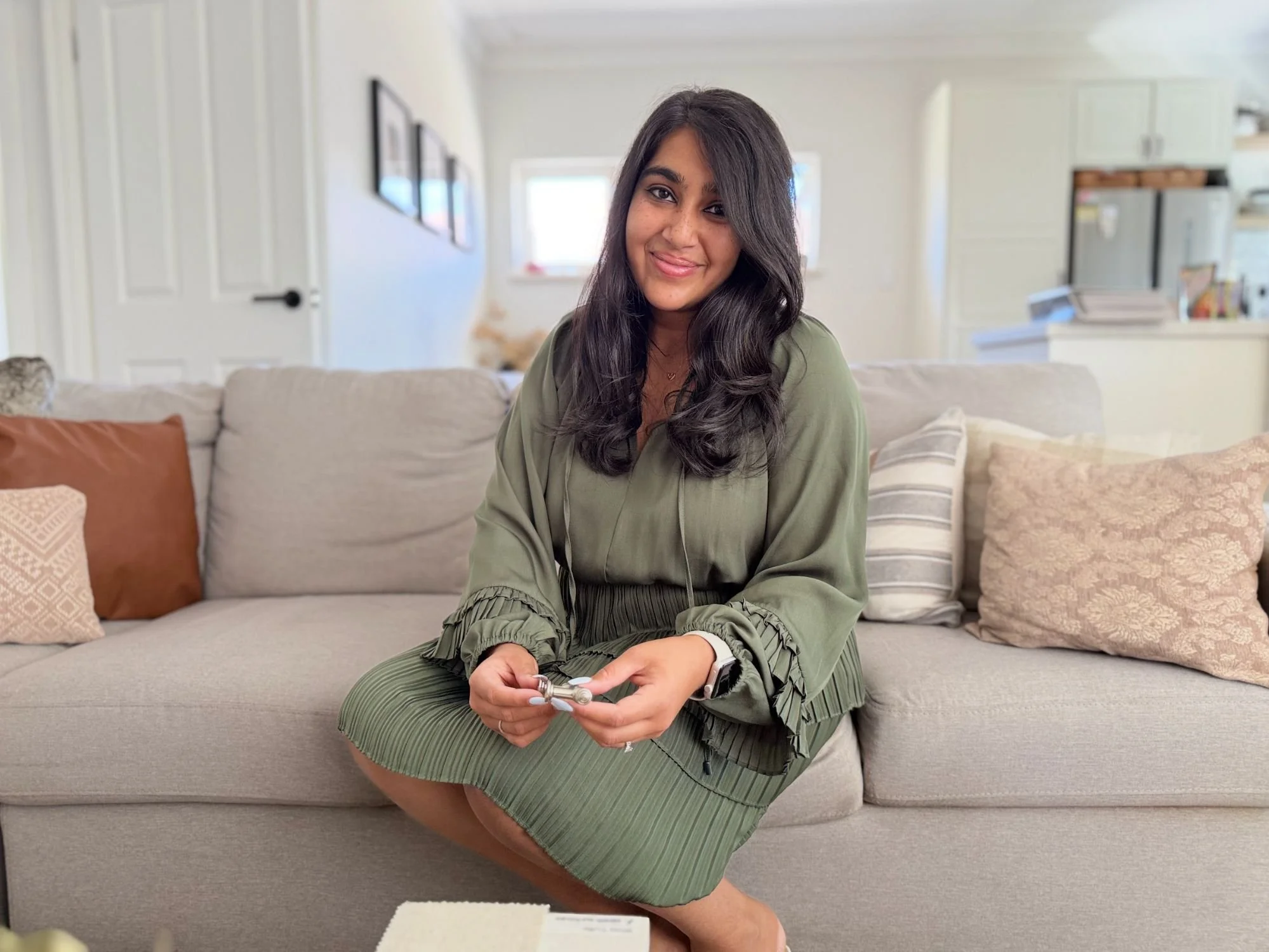 A young woman with long dark hair, smiling, seated on a beige couch in a well-lit living room. She is wearing a green outfit and holding a remote control.