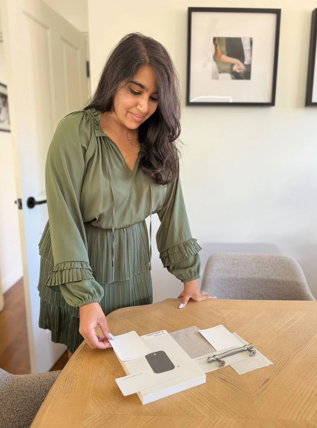 A woman in a green dress is leaning over a wooden table, examining a boxed electronic device, with a skateboard and some cloth on the table, in a room with framed artwork on the white walls.
