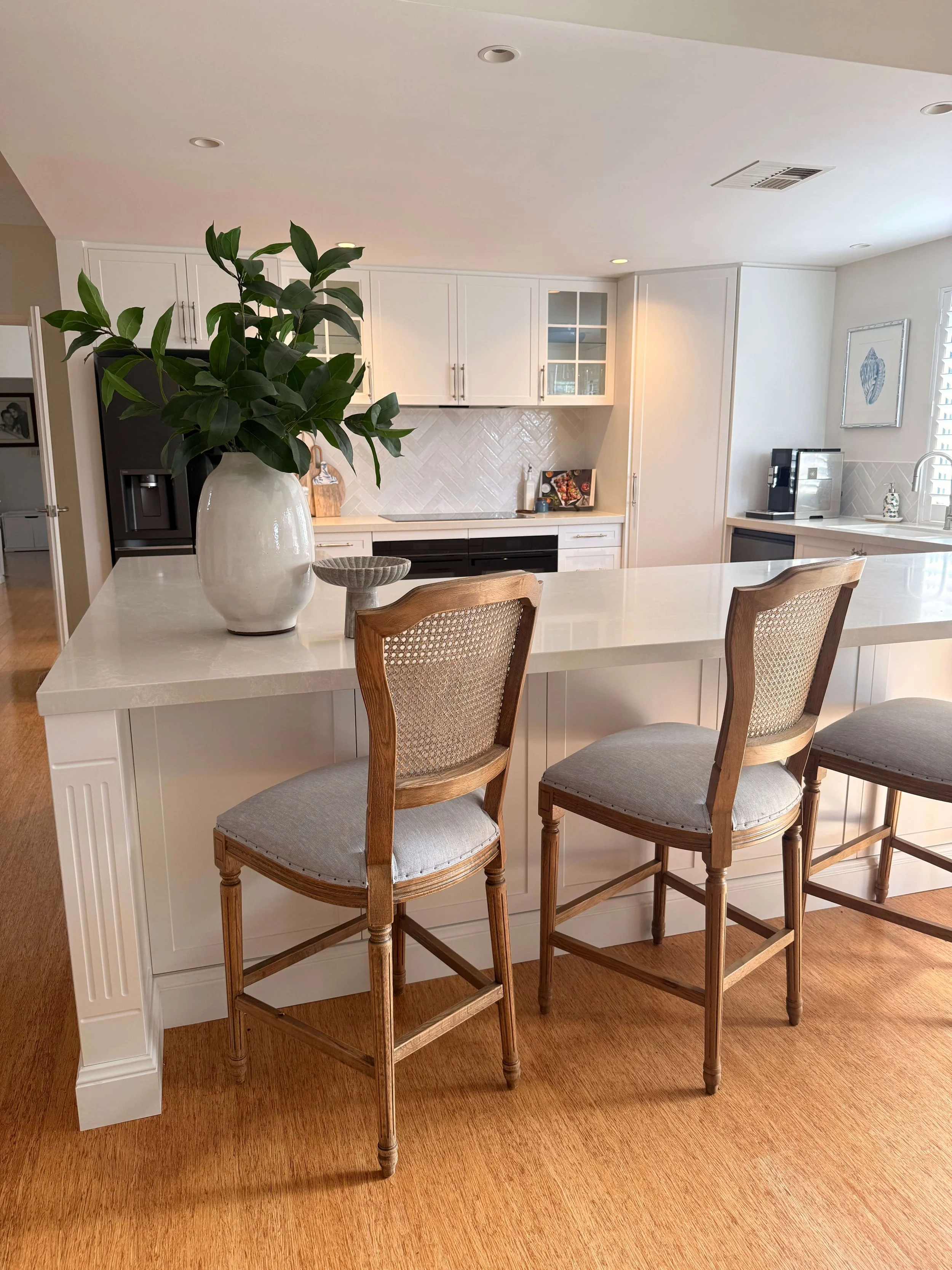 A kitchen with white cabinets, a large white island, a potted plant on the island, three wooden chairs with upholstered seats, a black refrigerator, and a coffee maker on the counter.