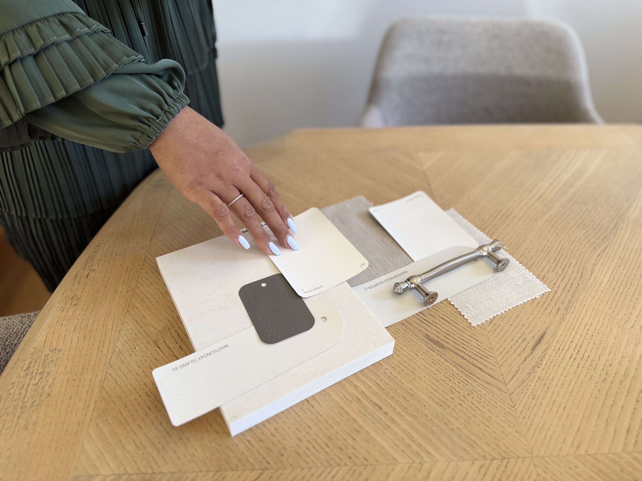 A person's hand with a wedding ring on the ring finger resting on a sample book of paint colors on a wooden table. There are color swatches and paint tool samples, and a gray upholstered chair in the background.