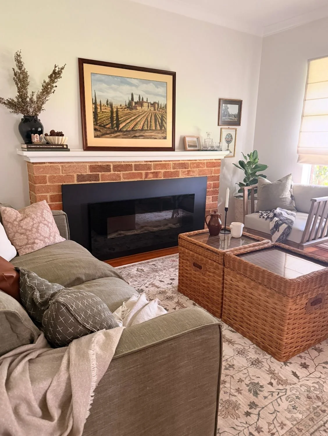 Living room with a brick fireplace, framed landscape painting, beige couch with decorative pillows, wicker coffee table, and a white armchair near a window with beige blinds.