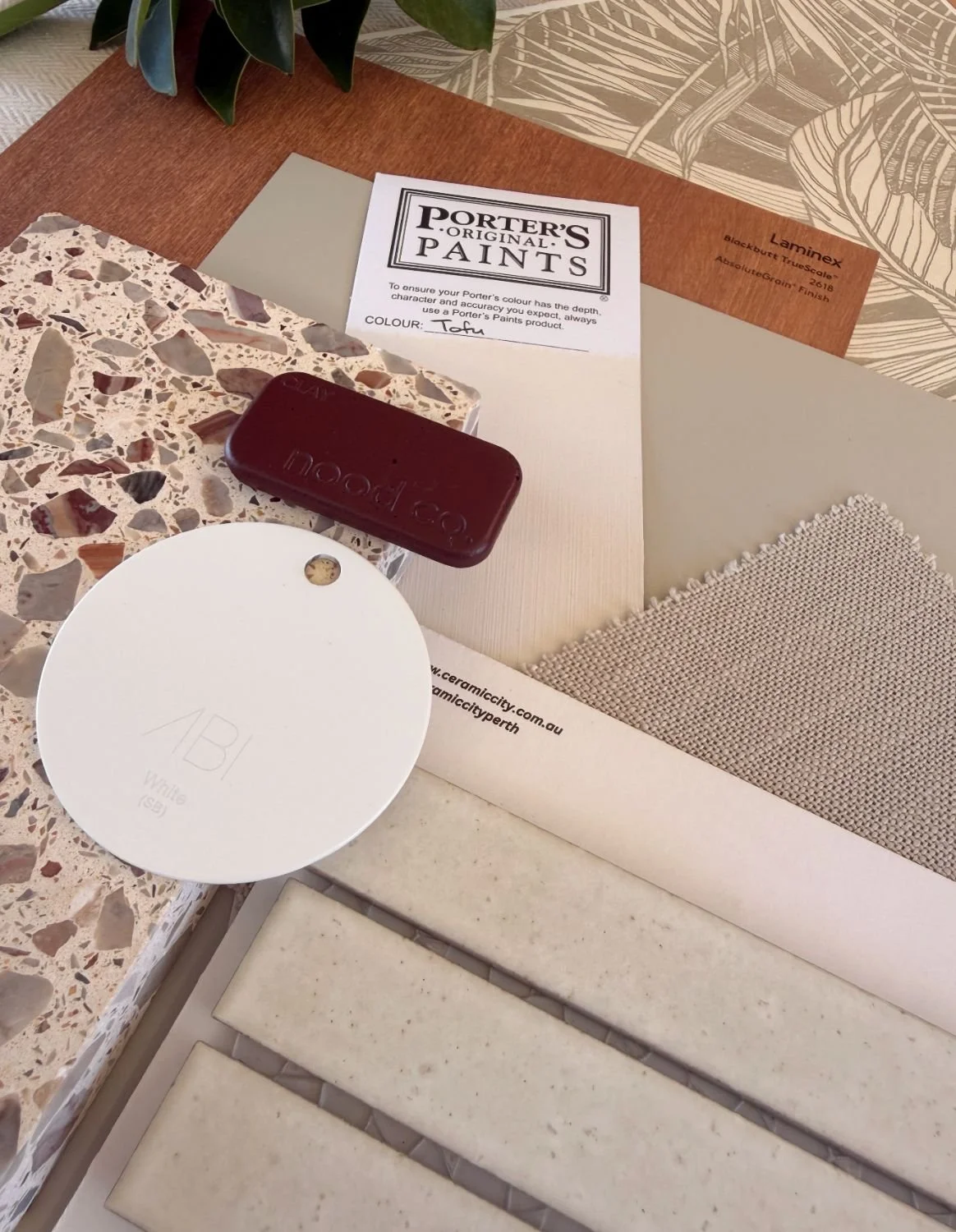 A collection of interior design and paint sample items including a Porter’s original paints sample card, a bar of chocolate, a terrazzo tile sample, a ceramic tile sample, a textured fabric swatch, and a round white ceramic tile with the letters 'ABI' and 'White' written on it, arranged on a work surface with a plant partially visible in the background.