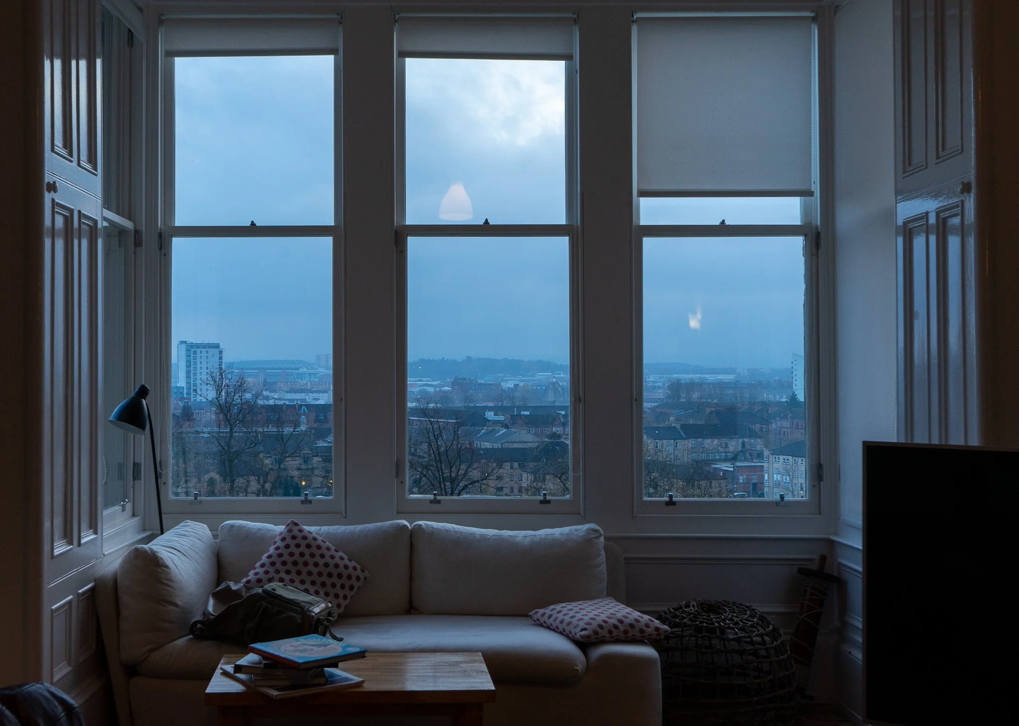 A living room with large windows showing an urban cityscape, a beige sofa with throw pillows, a wooden coffee table with books, a black floor lamp, and a flat-screen TV on the right.