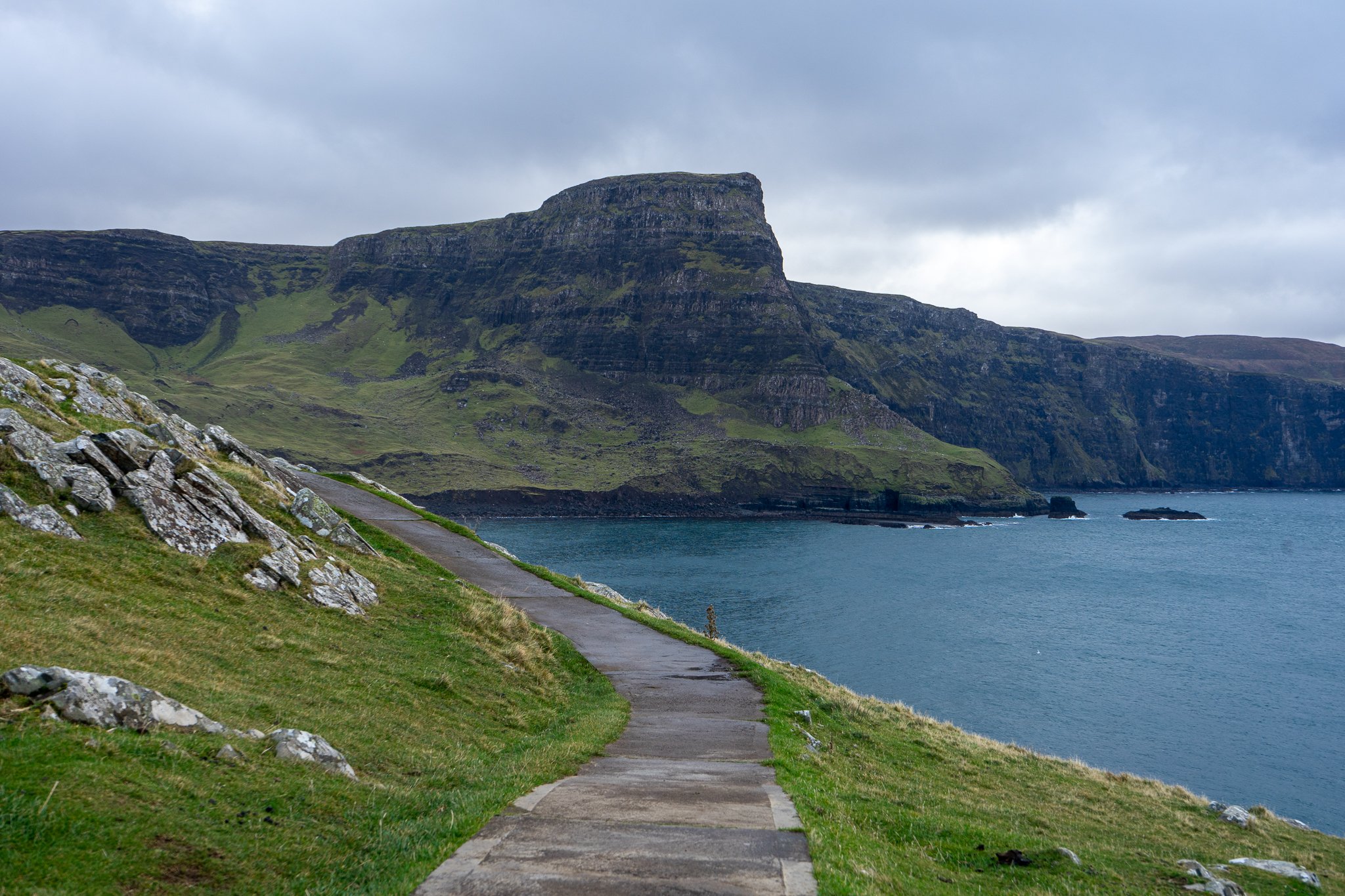 A winding concrete path along grass-covered hills leading to a rocky cliffside overlooking the sea with a cloudy sky overhead.