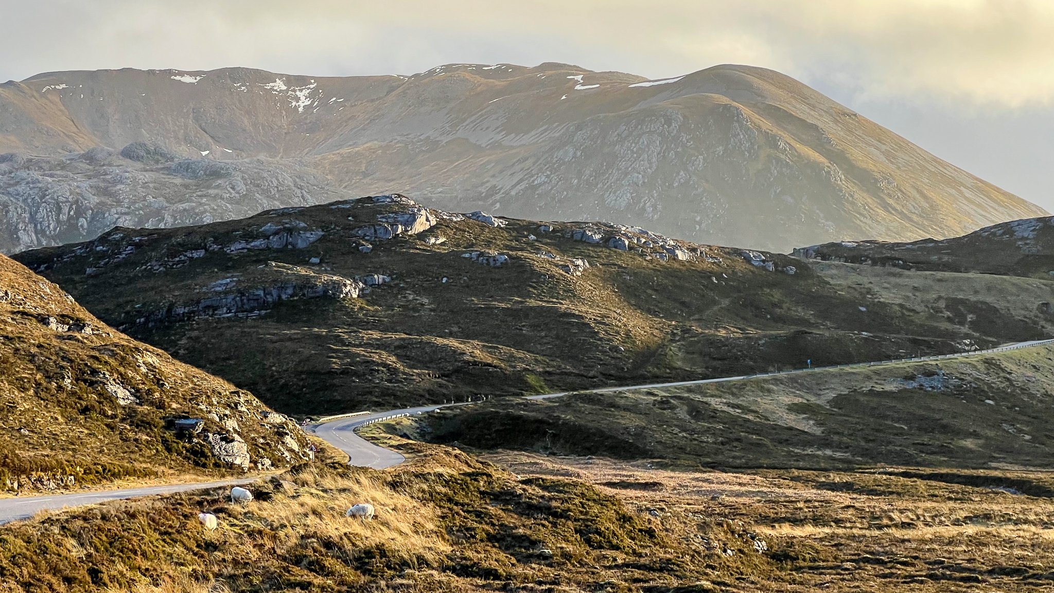 A winding mountain road in a hilly landscape with grass, rocks, and mountains in the background.