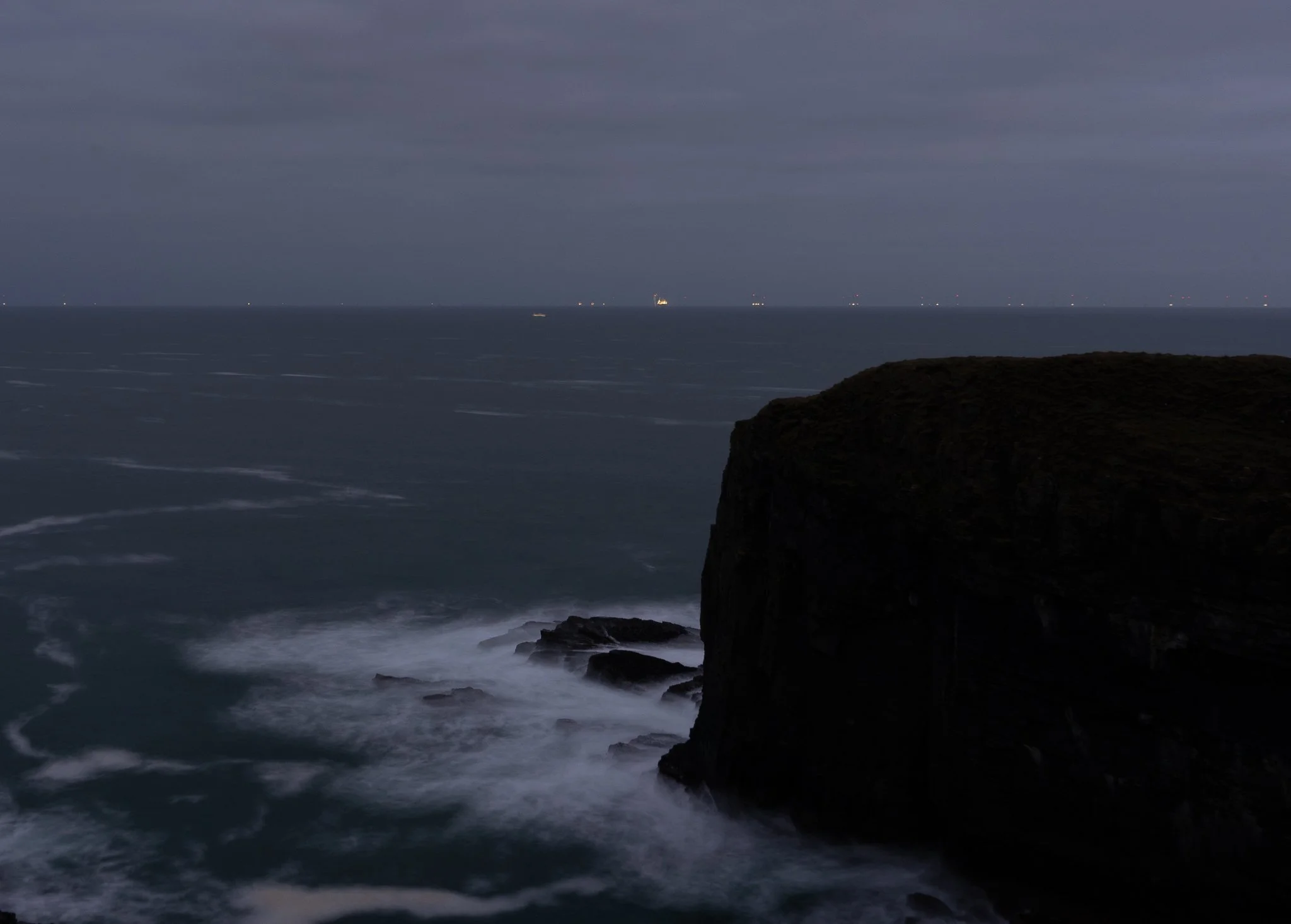 Dark ocean view with a cliff on the right and ships in the distance on a cloudy night