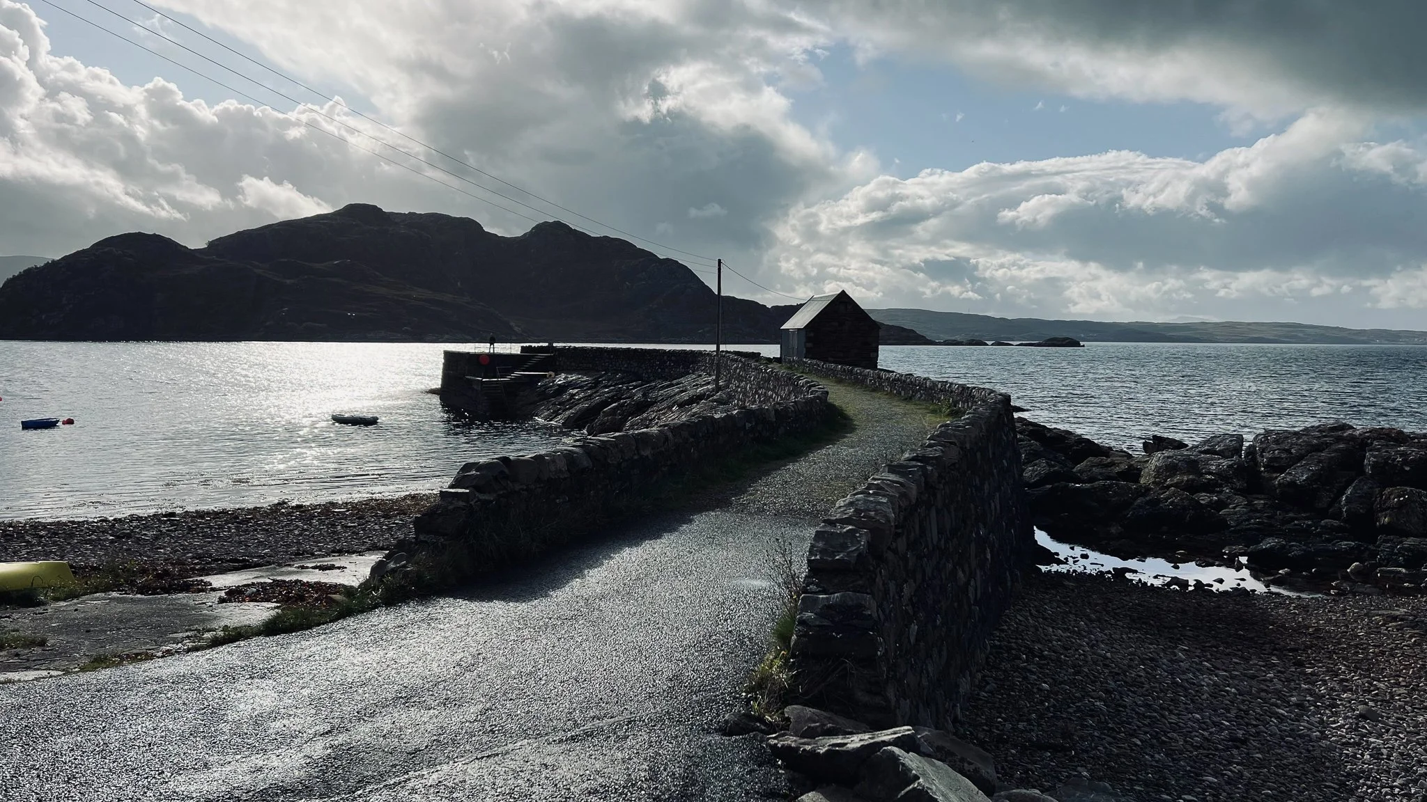 A stone pathway leading to a small wooden building located on a rocky shoreline, with calm water and mountainous landscape in the background under a partly cloudy sky.