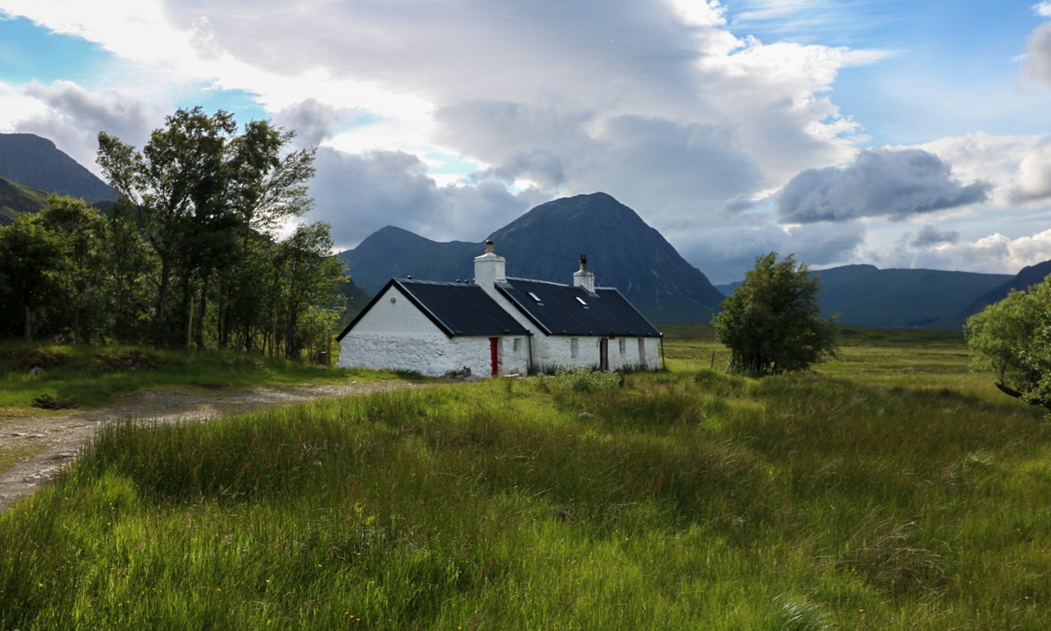 A white cottage with a black roof in a green field with mountains and cloudy sky in the background.