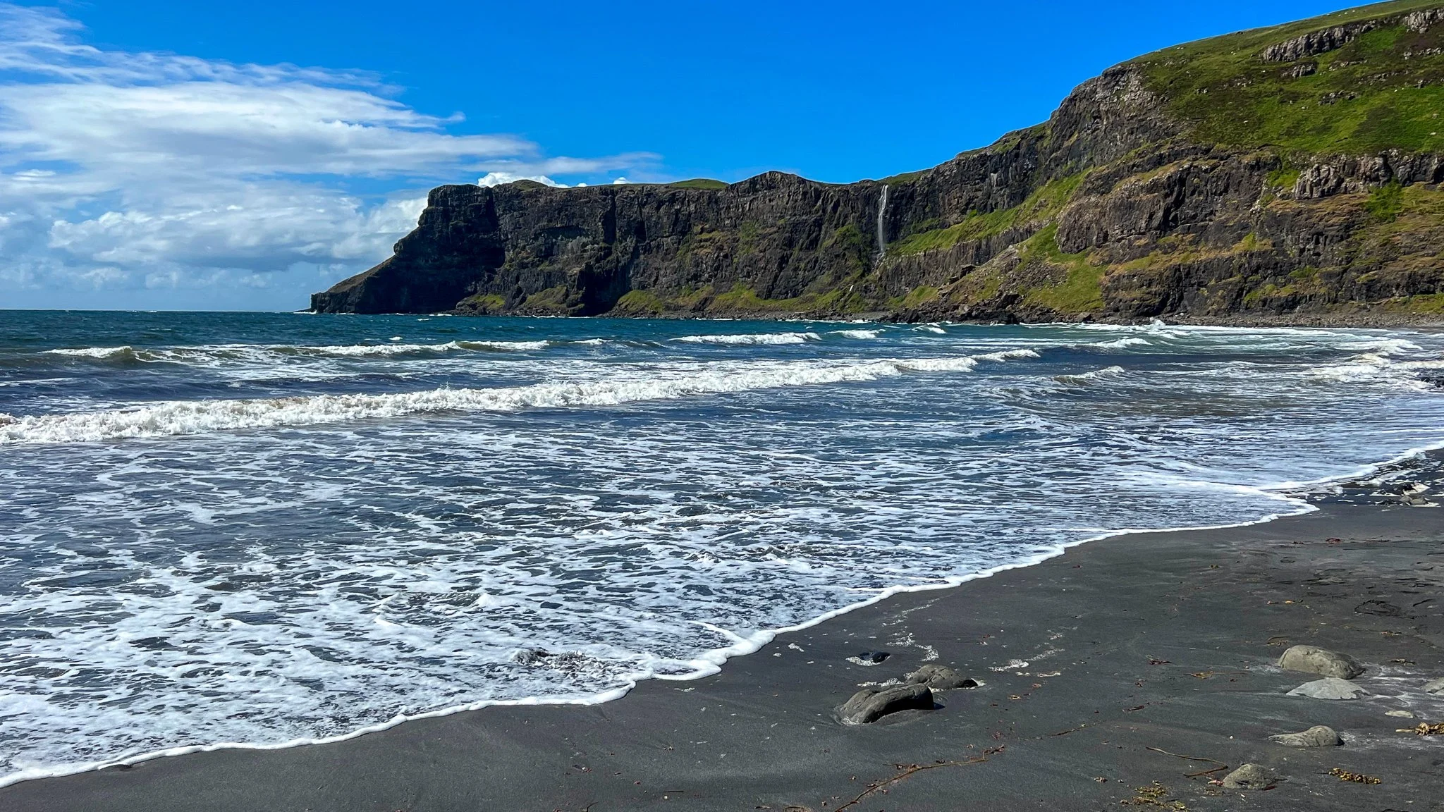 A scenic view of a beach with dark sand, ocean waves, rocky cliffs, green vegetation, blue sky with clouds, and a small waterfall cascading down the cliffs.