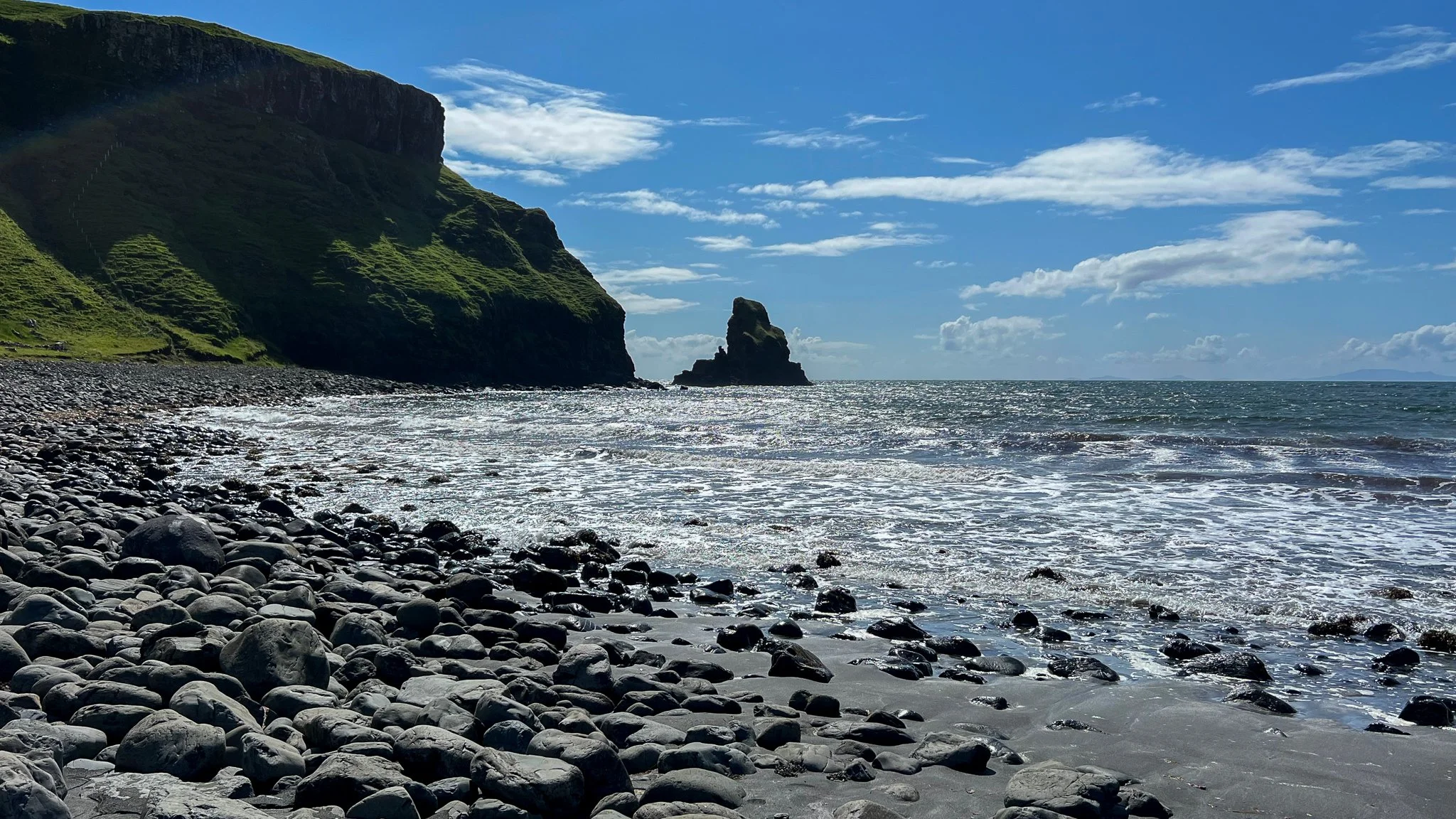 Rocky beach with pebbles, ocean waves, green cliffs, and a partly cloudy sky.