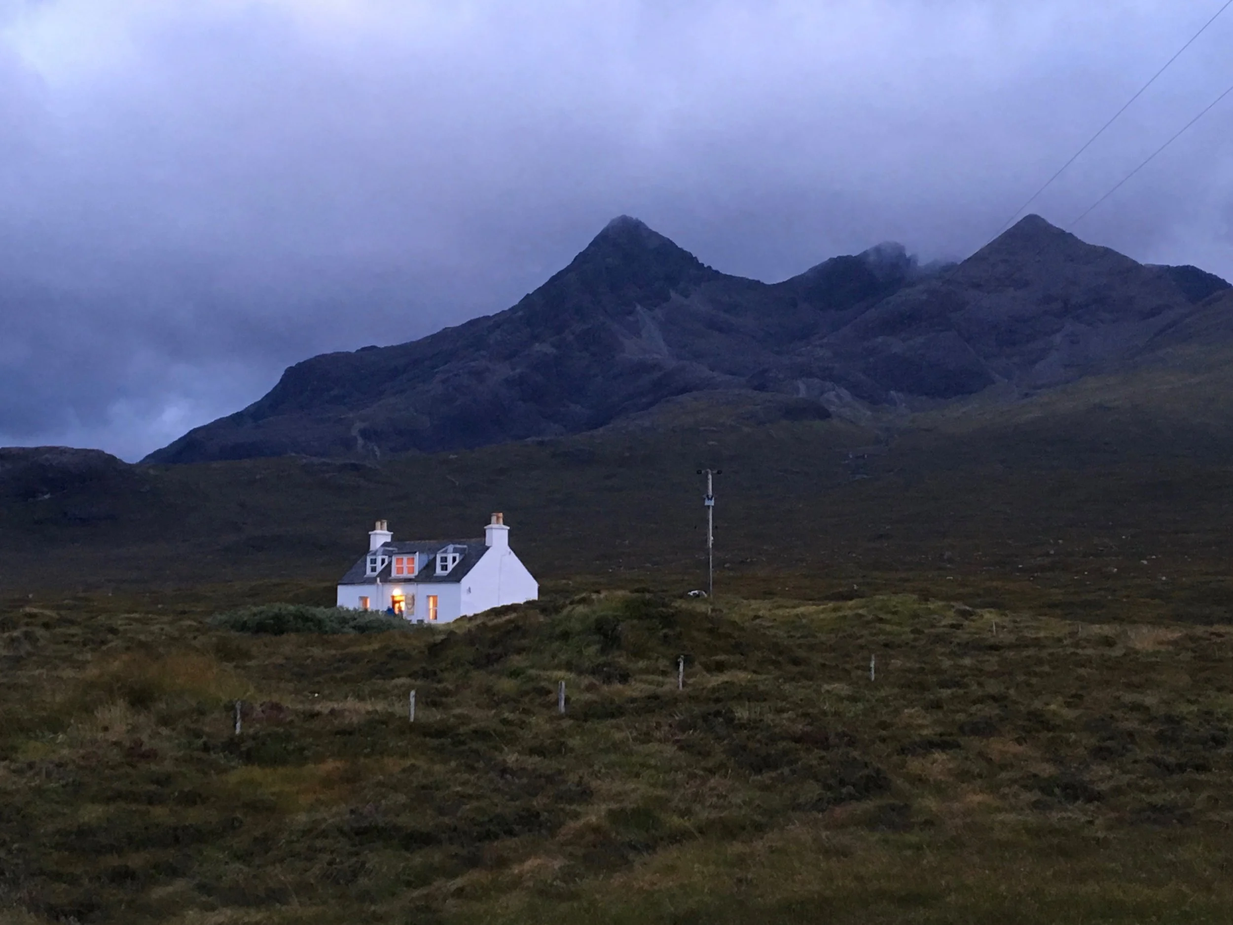 A white house or cottage with lit windows situated in a grassy moorland, with mountains in the background under a cloudy sky.