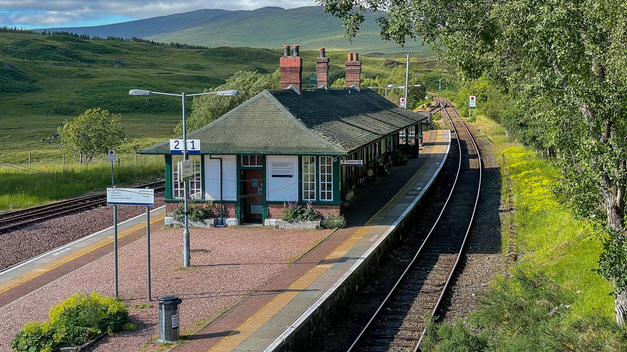 A small rural train station with green and white buildings, train tracks, and a scenic landscape of rolling green hills and mountains in the background.