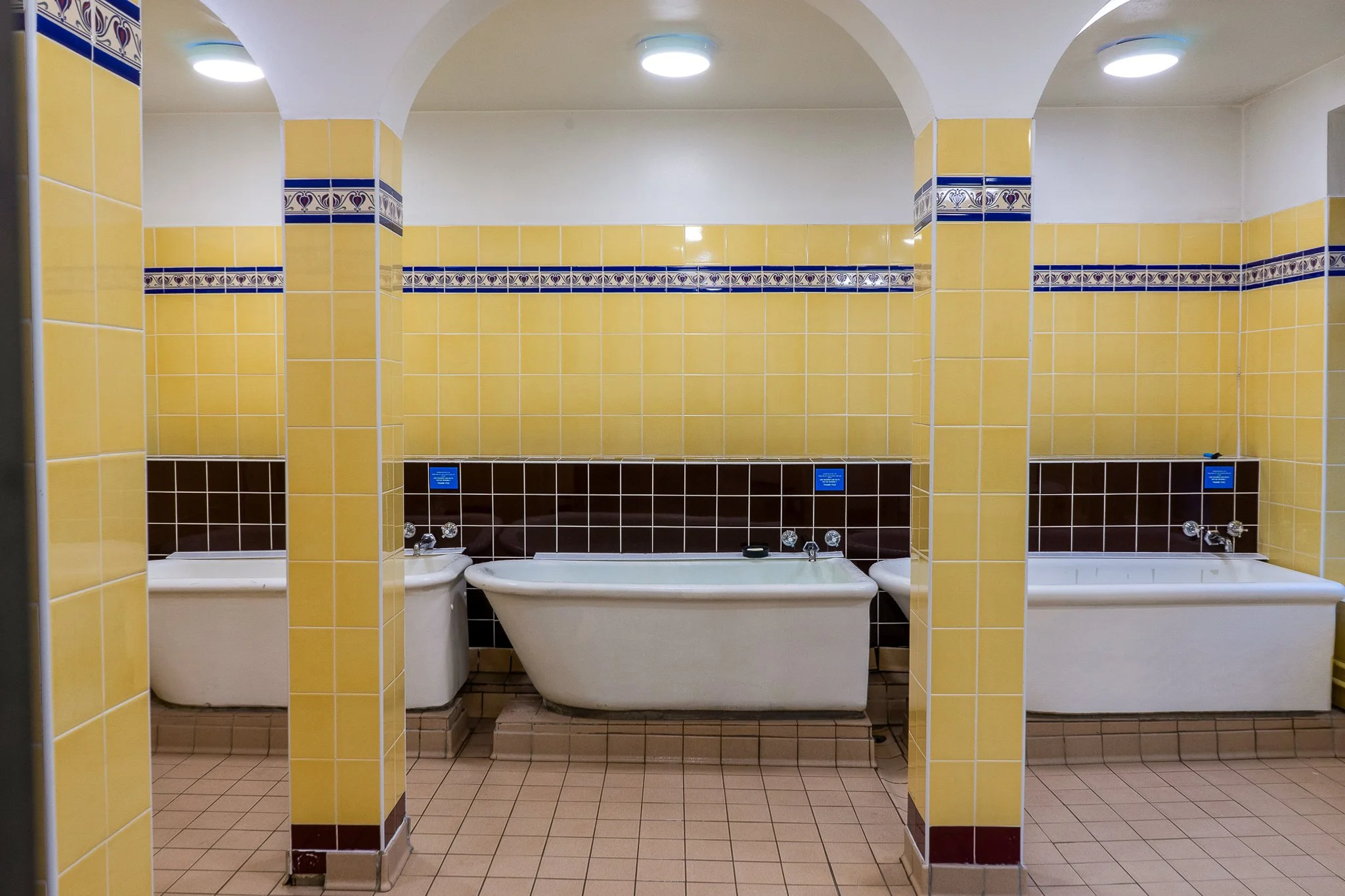 Public bathhouse with three vintage bathtubs inside yellow-tiled walls and brown accents, lit by ceiling lights.