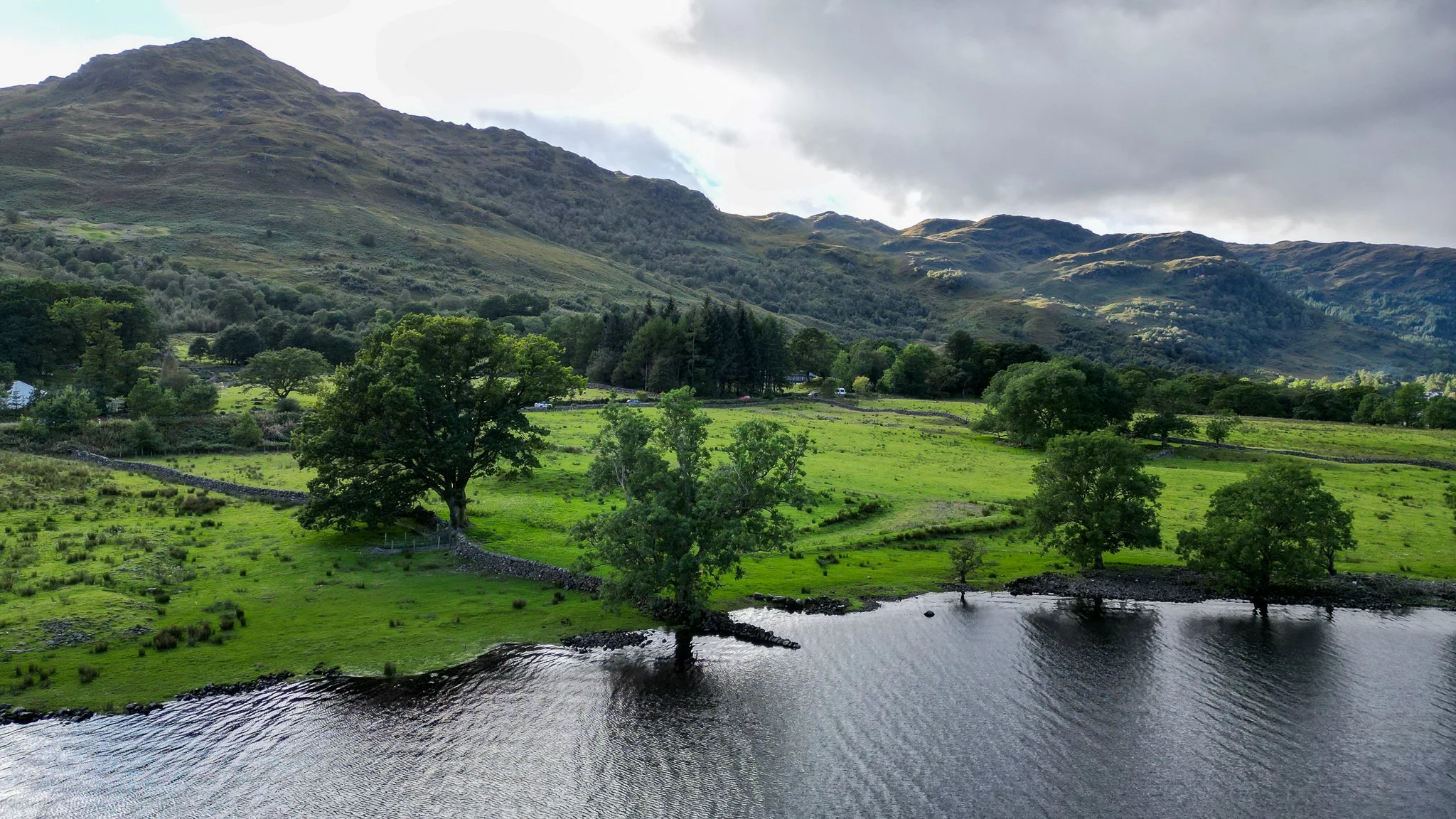 Scenic landscape with green fields, trees, and a body of water in the foreground, with mountains and cloudy sky in the background.
