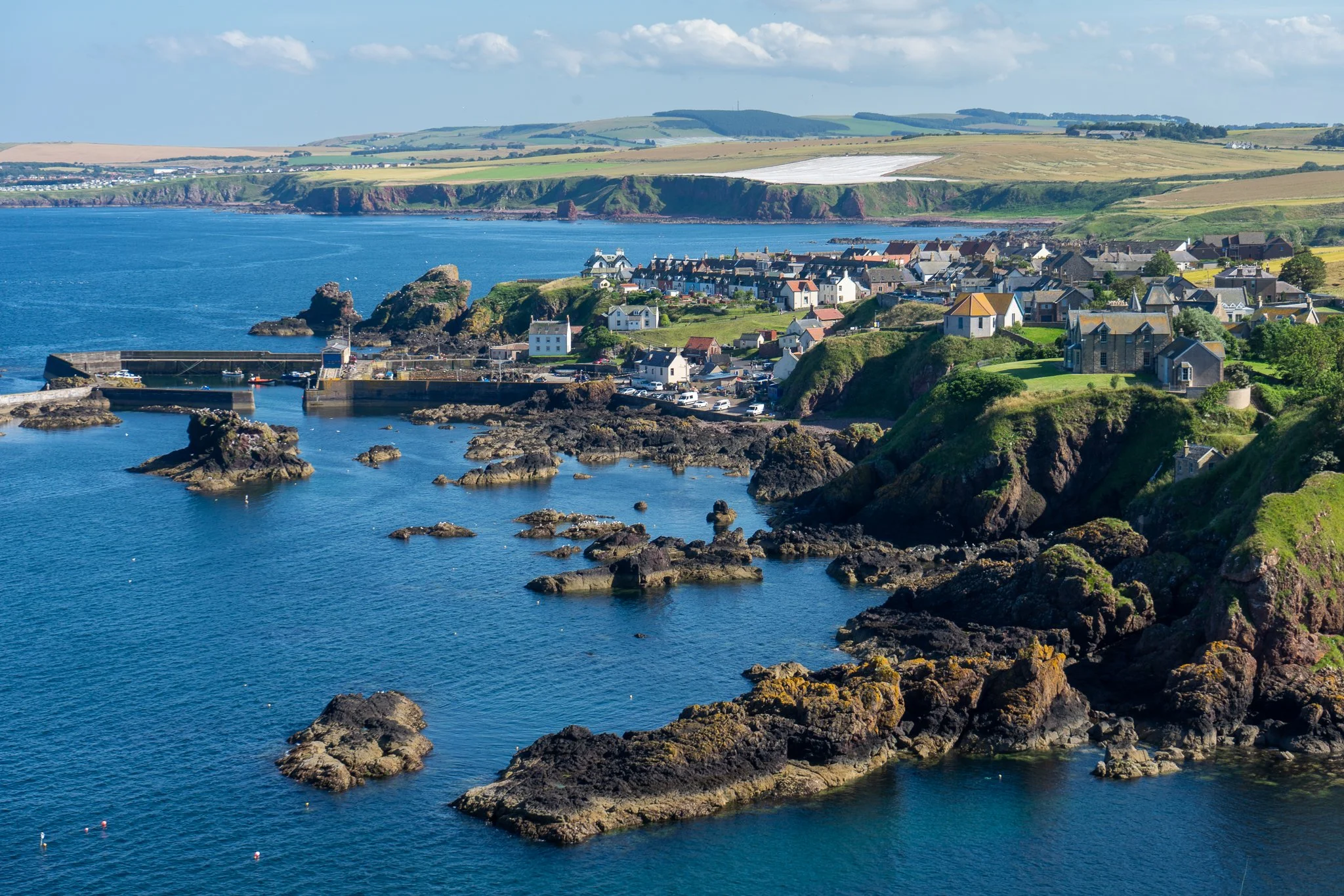 Coastal village with houses on cliffs, rocky shoreline, harbor with boats, and rolling green hills in the background under a partly cloudy sky.