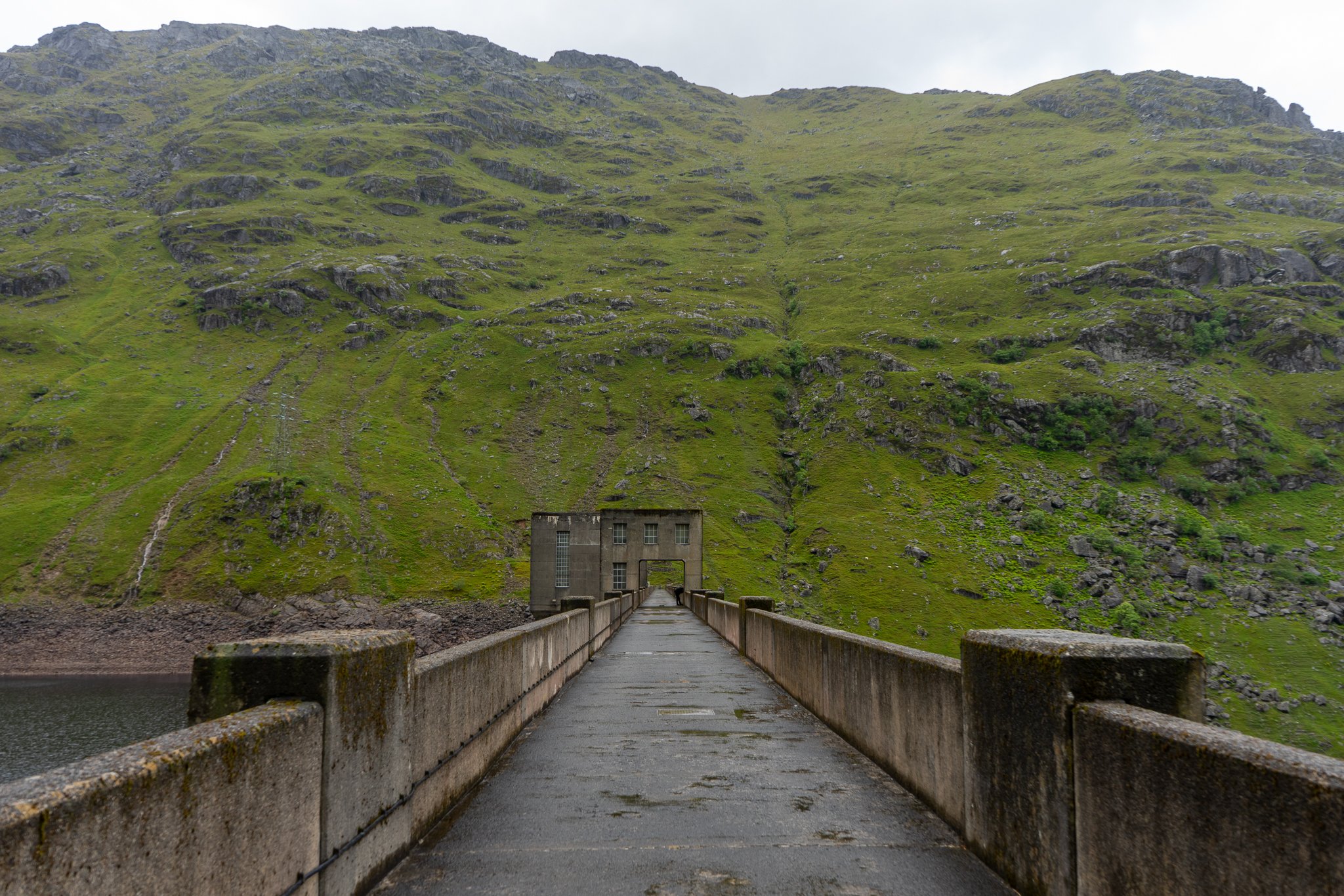 A concrete dam stretch across a green mountainous landscape, with a building at the end of the dam.