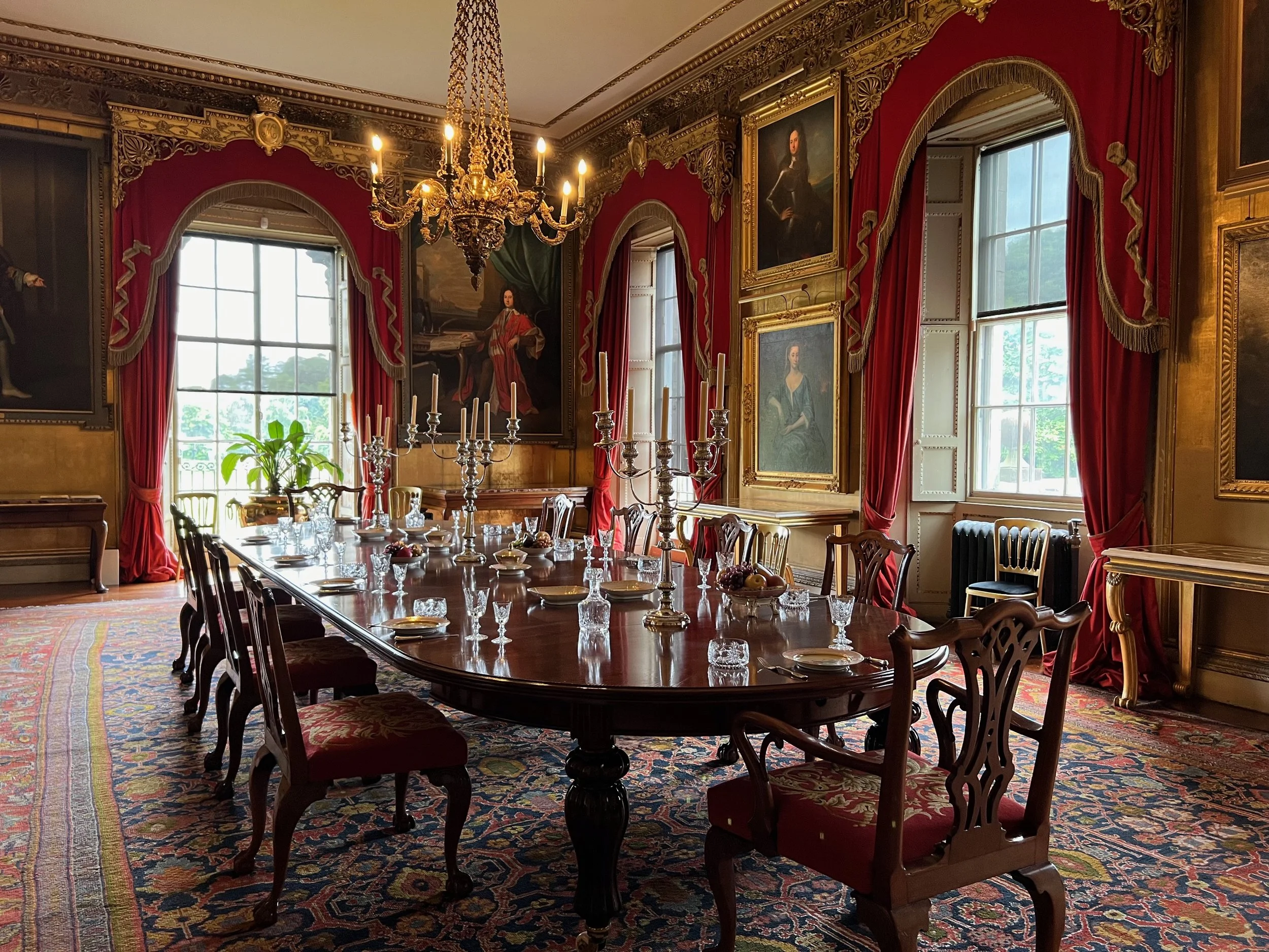 An opulent dining room with a long wooden table set with crystal glasses, silver candlesticks, and fruit bowls. The room features large windows with red velvet curtains, ornate gold trim, framed portraits, a chandelier, and a Persian rug on the floor
