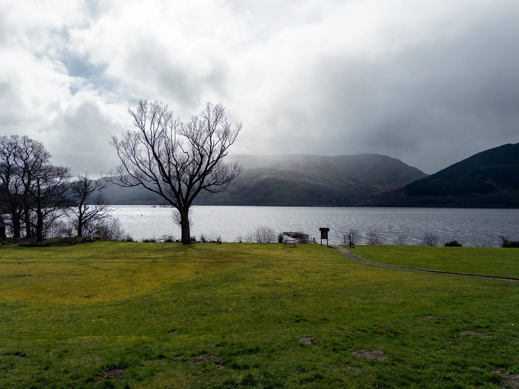 A park by a lake with leafless trees, green grass, benches, and a dock, with hills and cloudy sky in the background.