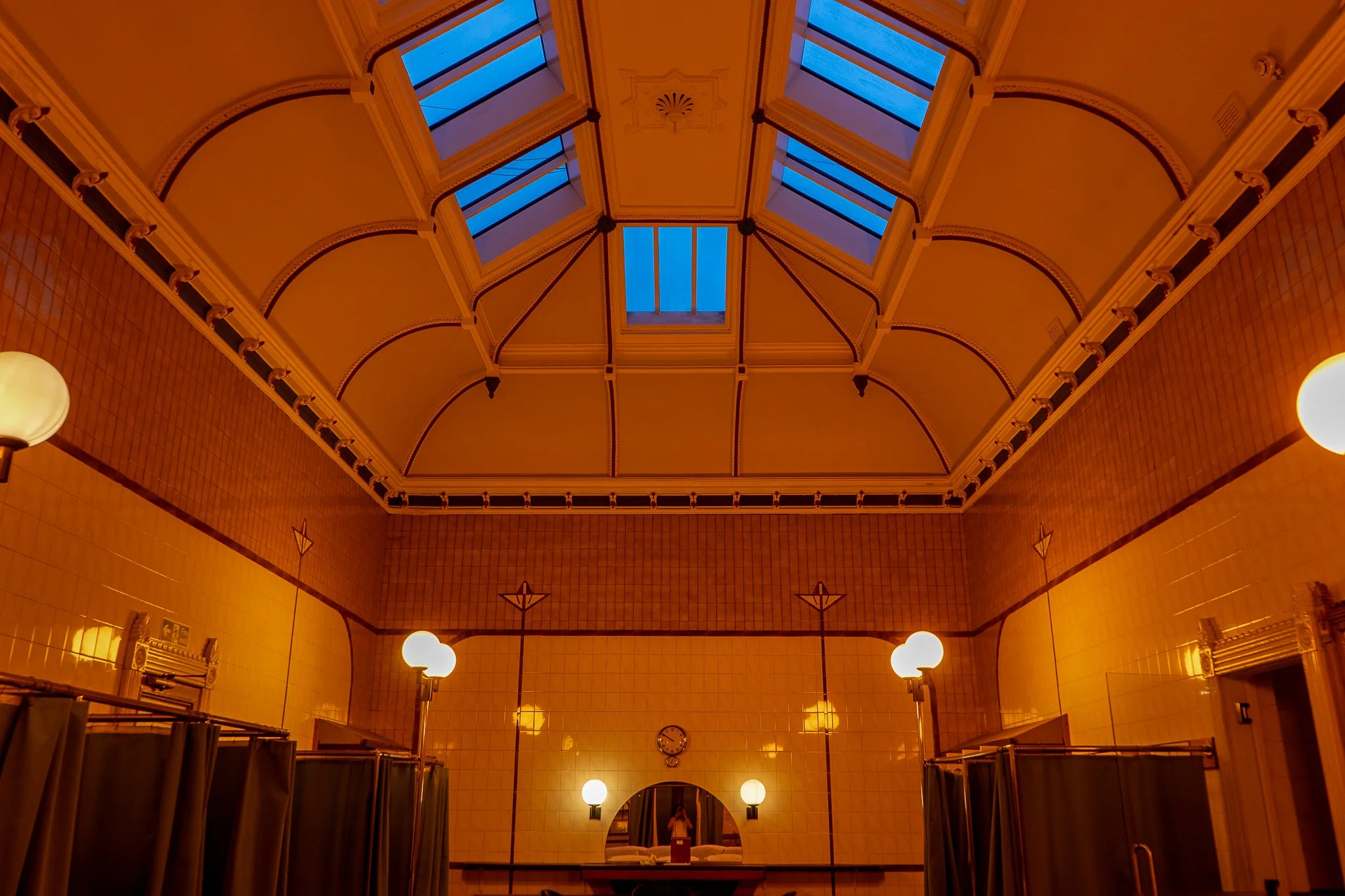 Interior of a tiled room with a high arched ceiling, large skylights, and warm lighting fixtures, including a clock and mirrors on the wall.