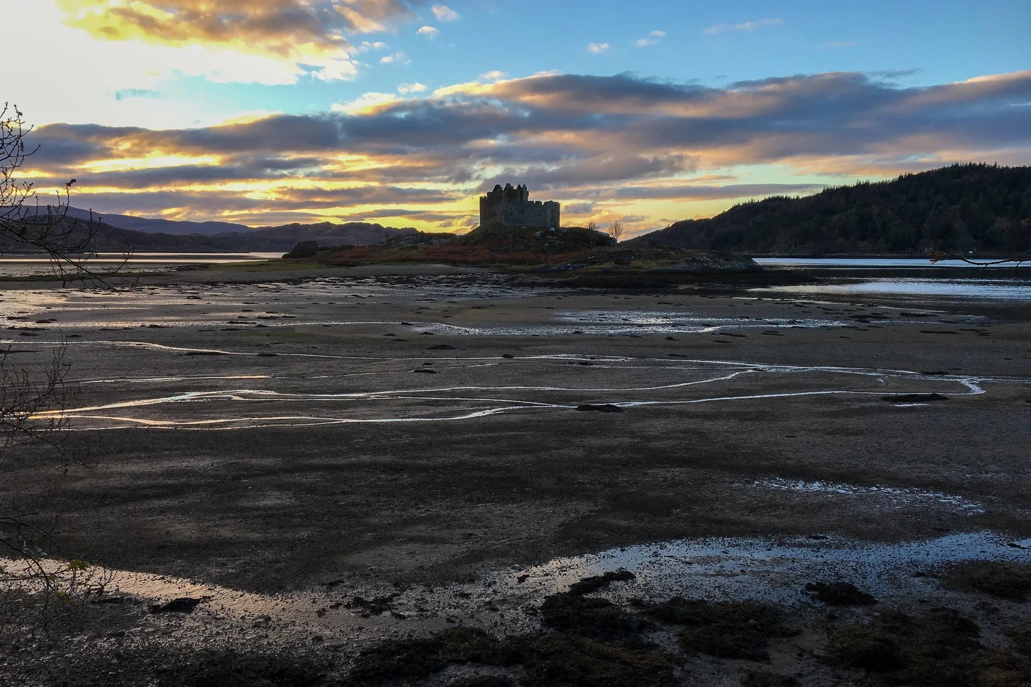 A scenic landscape with a castle in the distance, surrounded by water, hills, and a partly cloudy sky during sunset.