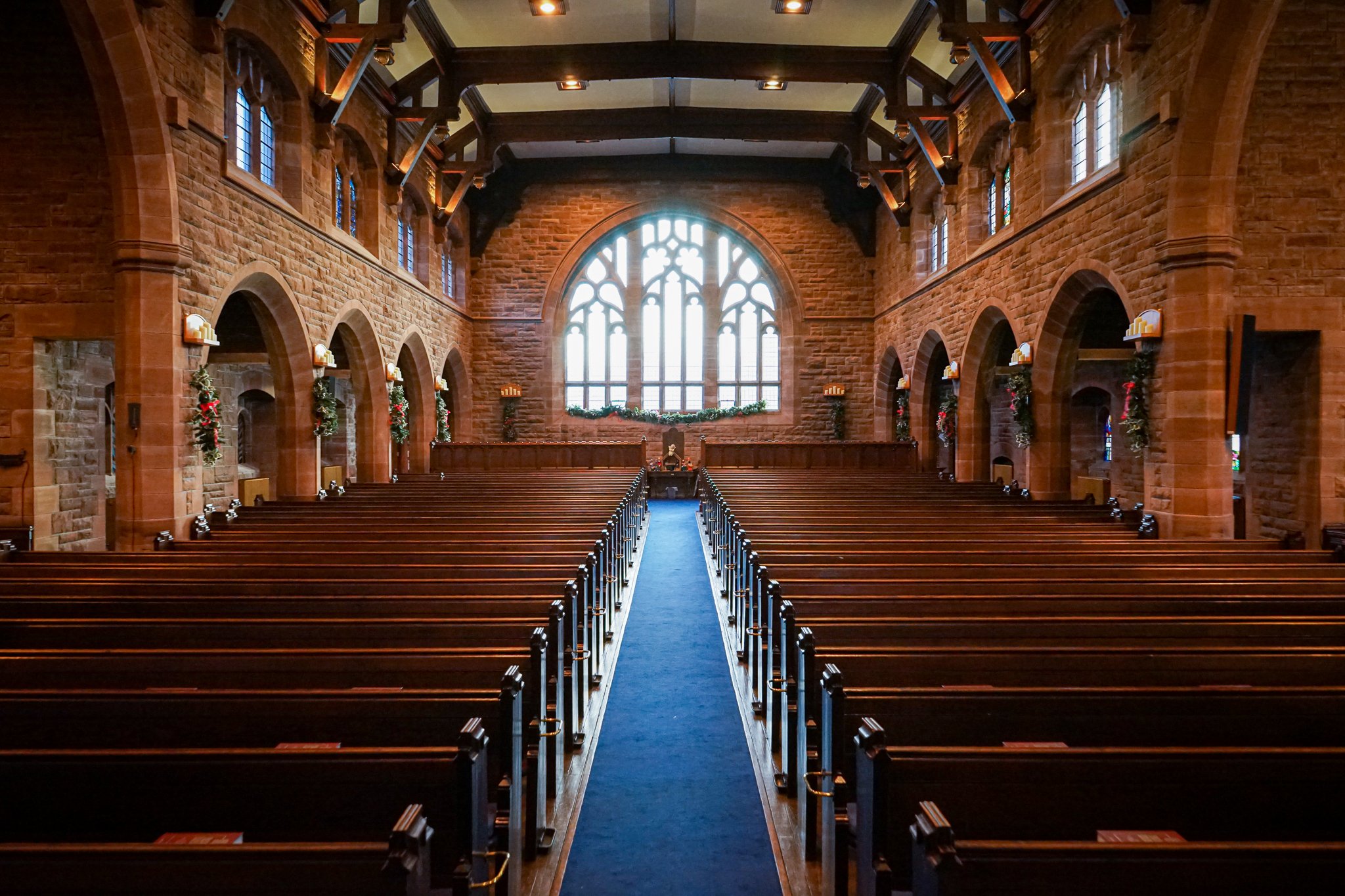 Interior of a church with wooden pews, arched windows, and Christmas decorations along the walls.