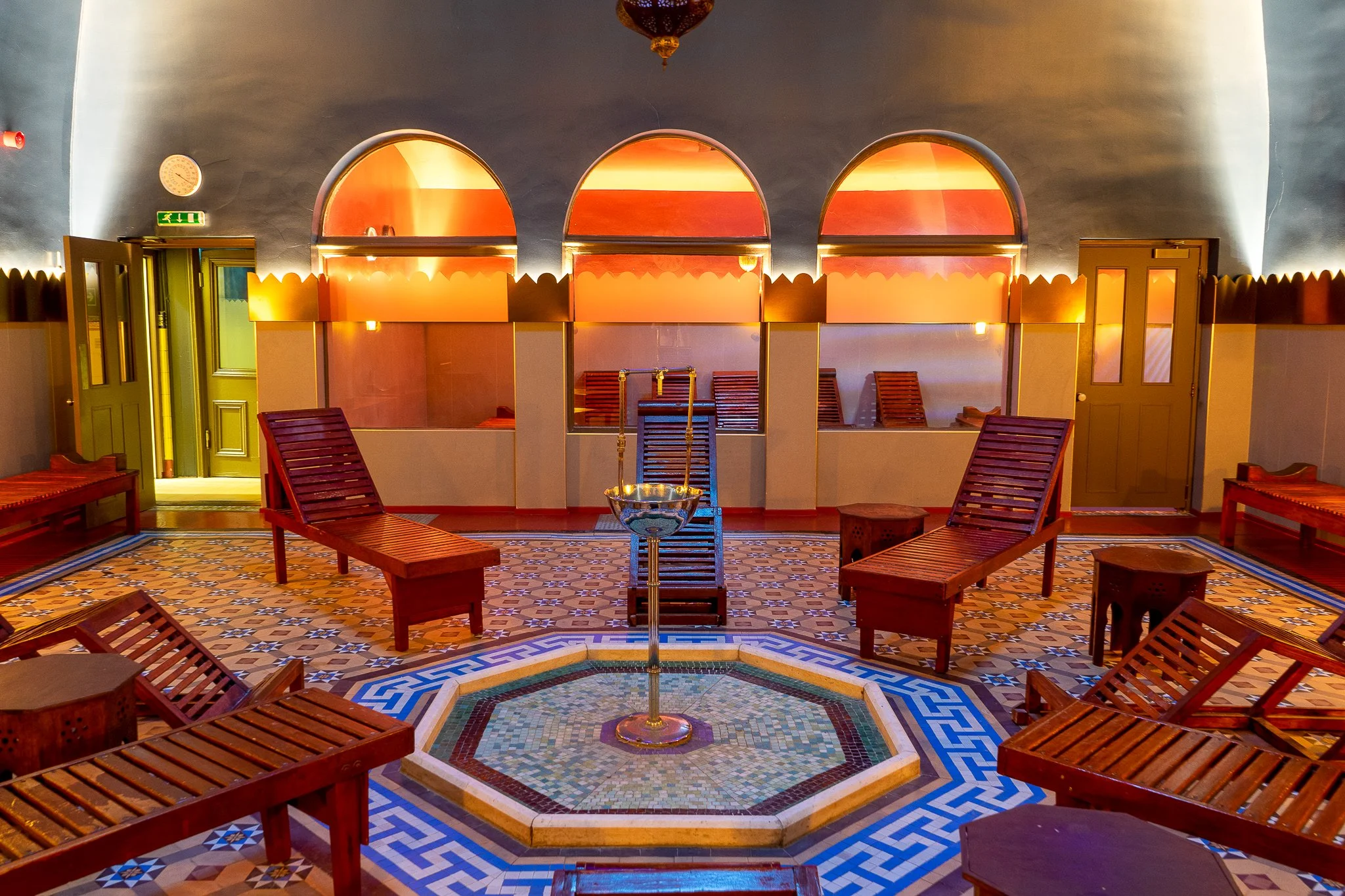 An indoor relaxation room with wooden lounge chairs arranged around a central small fountain. The room features a patterned tile floor, arched windows with warm lighting, and doors on either side.